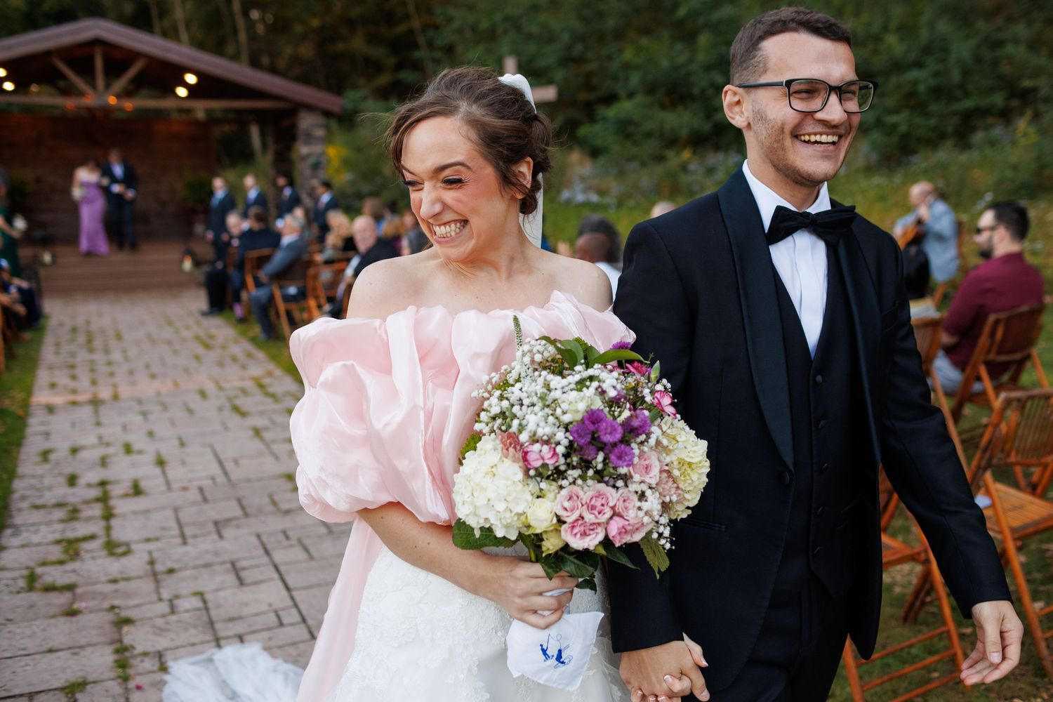Joyful newlyweds walk down aisle after ceremony, bride holding colorful bouquet in pink wedding dress.