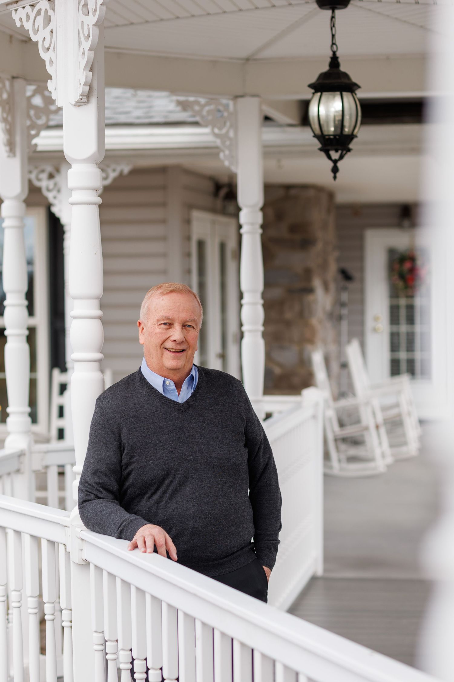 Person in a black sweater smiling while standing on a white Victorian-style porch with ornate columns and railings.