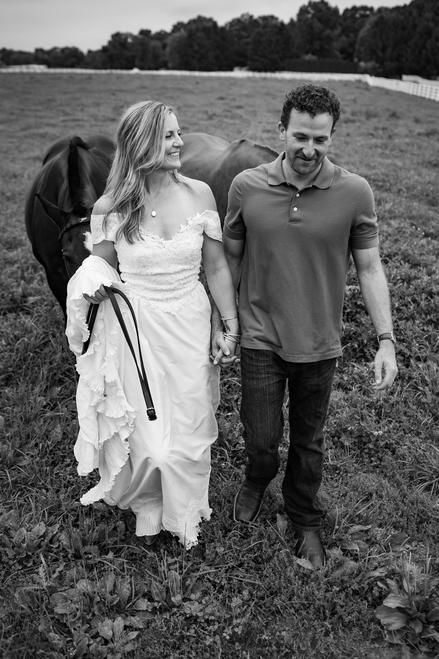 Black and white photo of a smiling couple walking hand in hand with a horse in a scenic pasture.