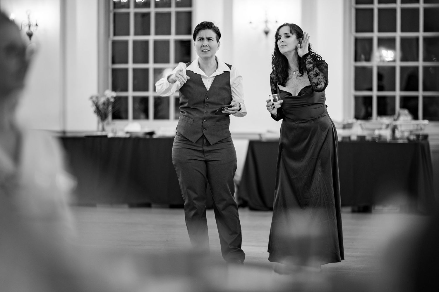 Elegant black and white photo of people in formal attire at an indoor venue with large windows.