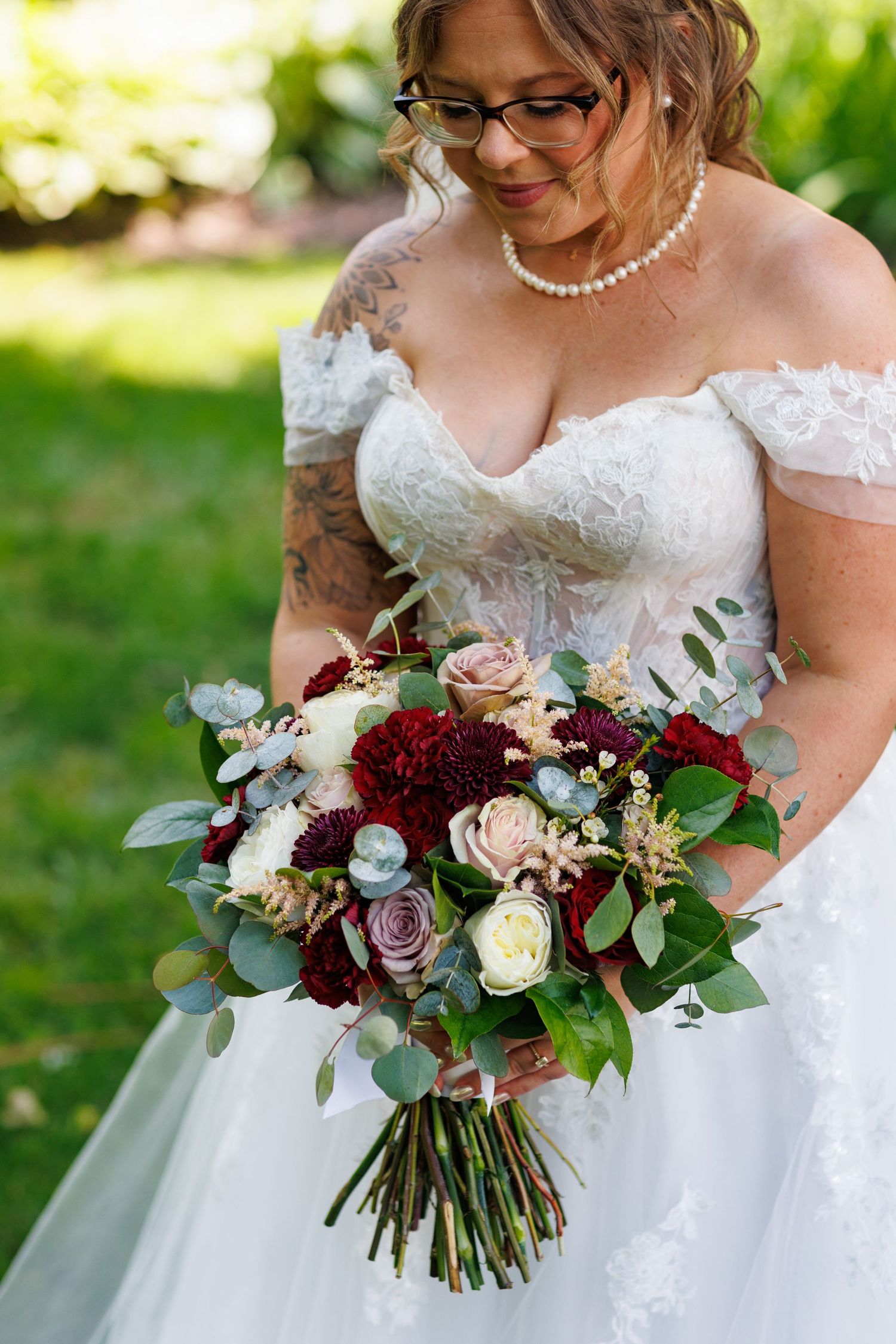 Bride in off-shoulder lace gown holding a lush burgundy and blush wedding bouquet with eucalyptus greenery outdoors.