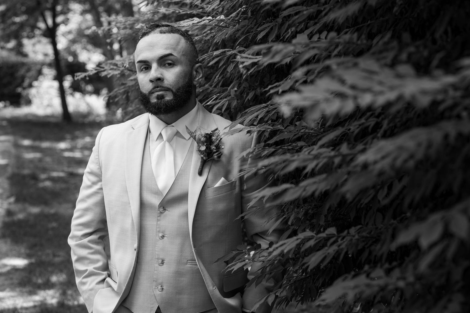 Dapper groom in light grey suit with boutonniere posing beside evergreen tree in black and white.