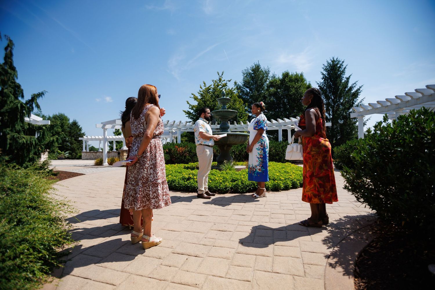 Four people socializing outdoors on a garden patio with lush greenery and a white pergola on a sunny day.