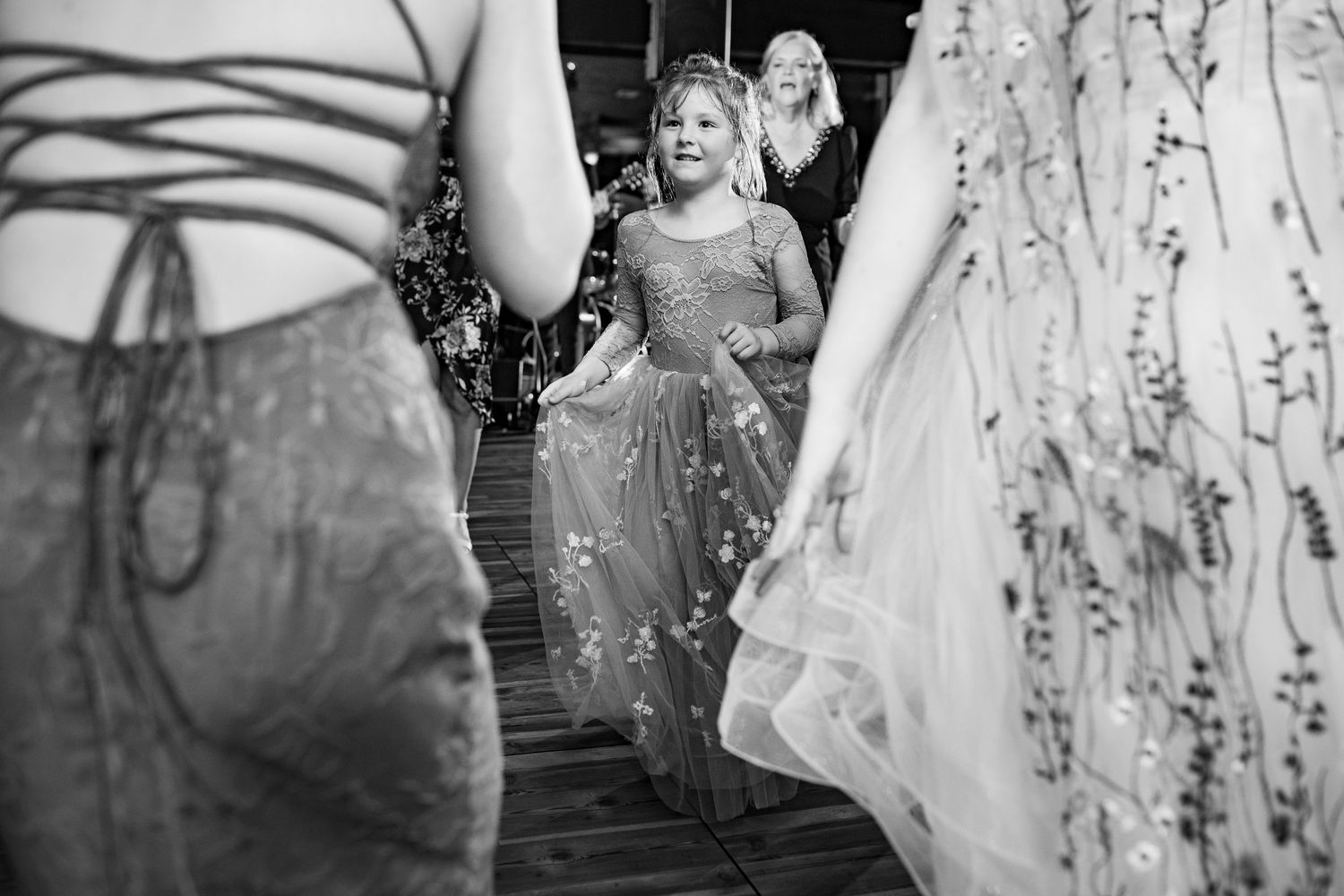 Excited flower girl twirls in sparkly dress among wedding guests on dance floor in black and white.