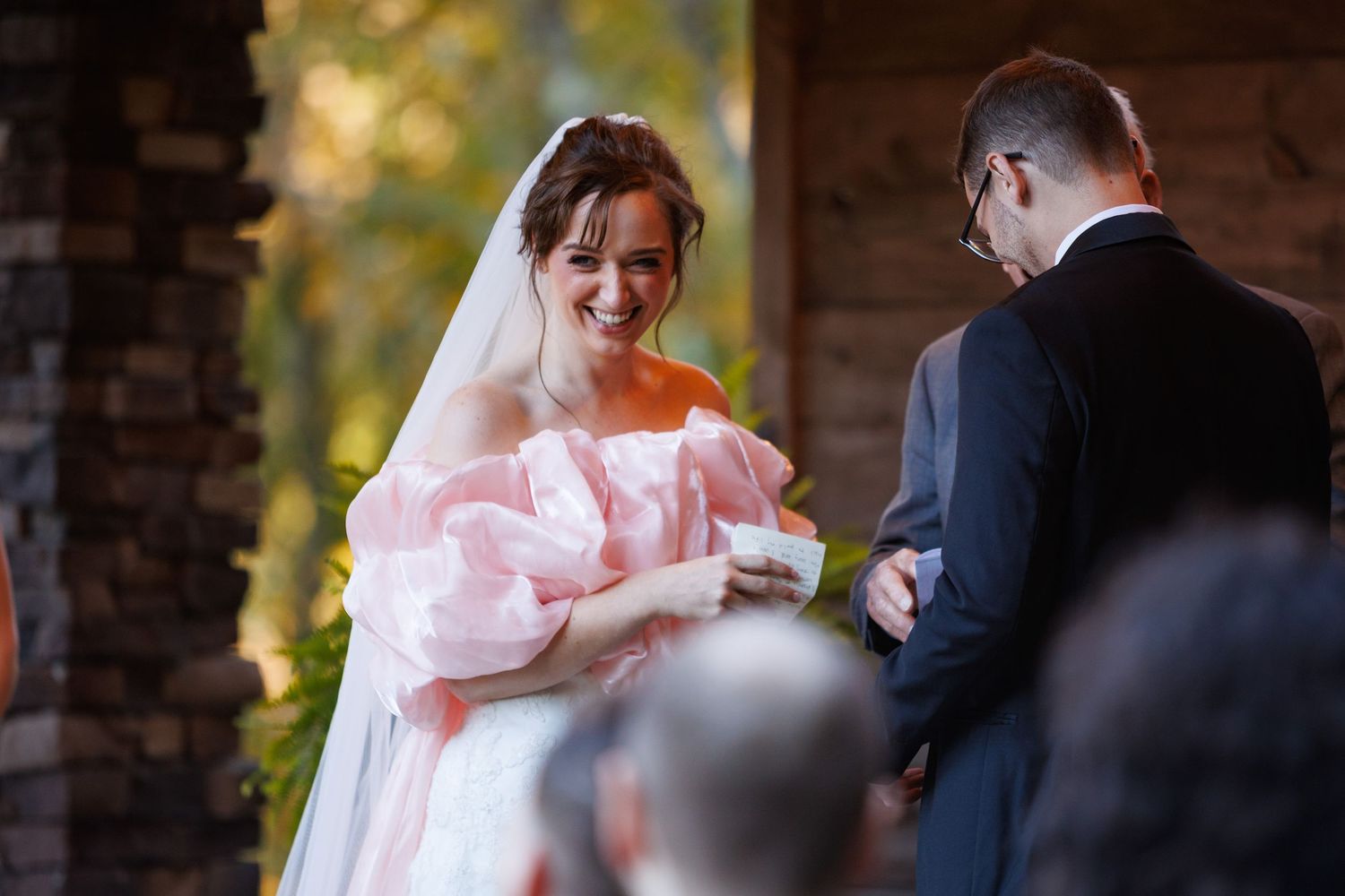 A bride in a pink and white gown smiles during wedding vows at a rustic outdoor ceremony.
