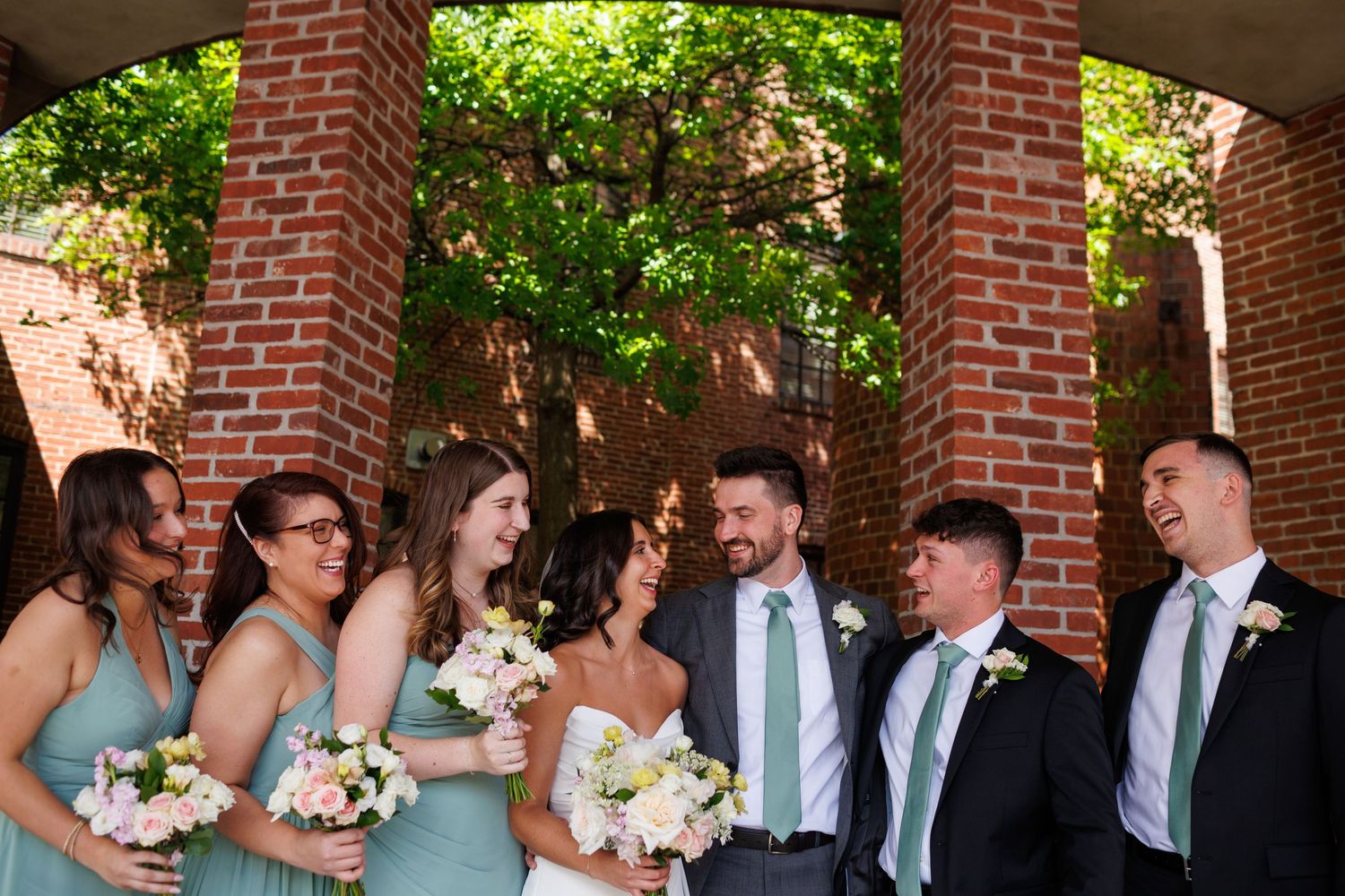 Wedding party laughing together outdoors near brick columns, bridesmaids in sage green dresses with white bouquets.