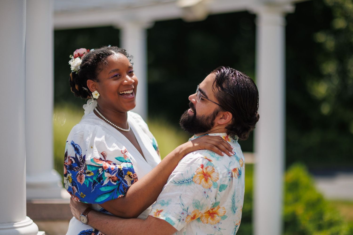 A smiling couple in floral shirts share a joyful embrace outdoors near white columns on a sunny day.