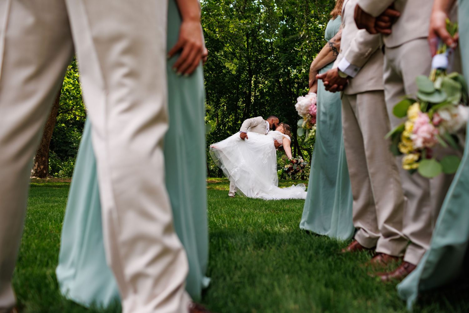 Groom dipping bride for a kiss between bridesmaids and groomsmen on green lawn.