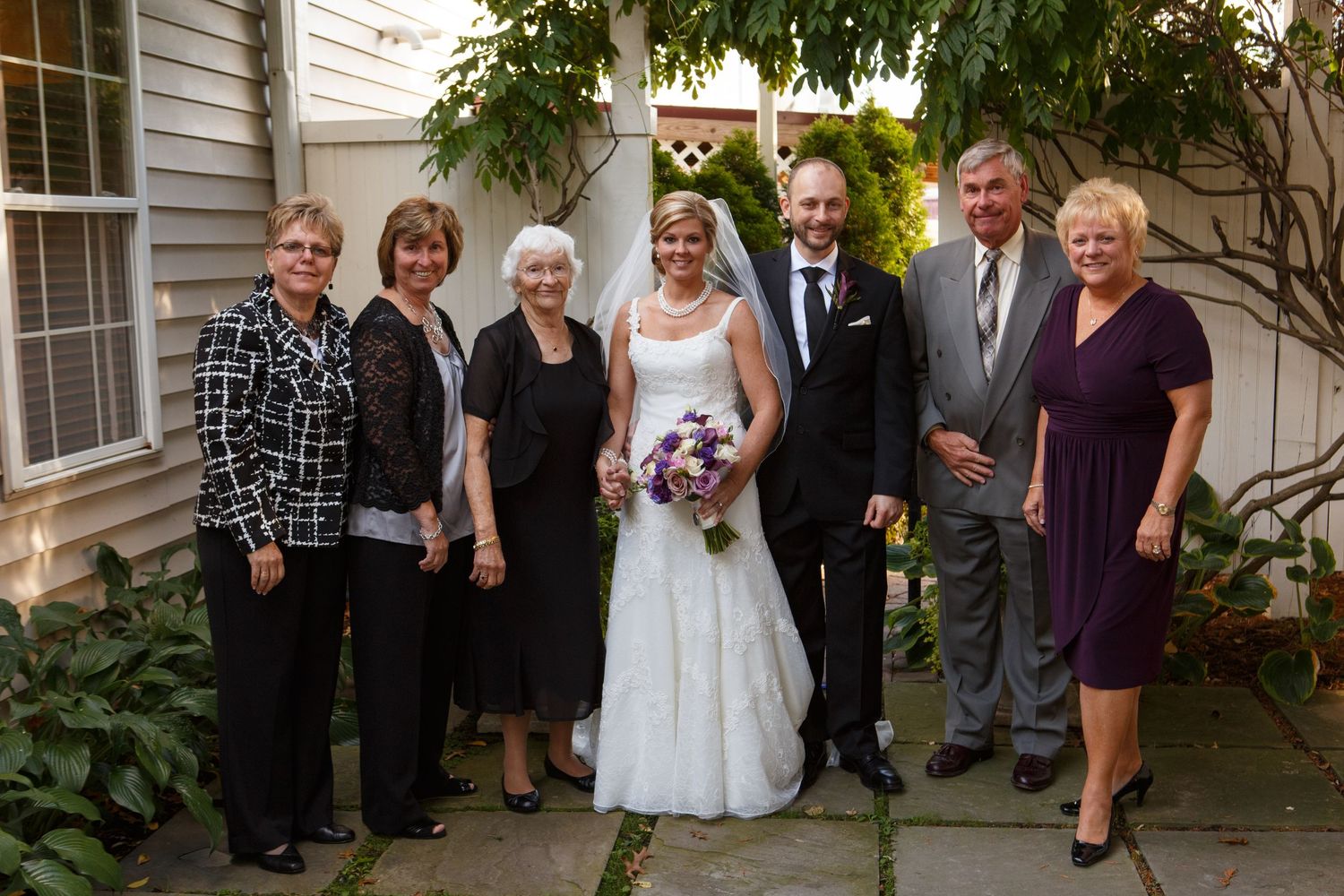 A group of formally dressed people pose together outdoors against a white building with greenery and purple flowers.