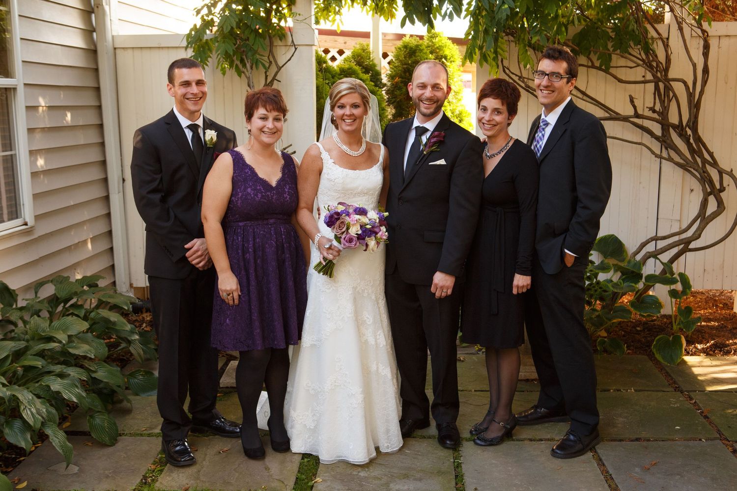 A wedding party stands together in formal attire in front of a vine-covered wall on a bright sunny day.