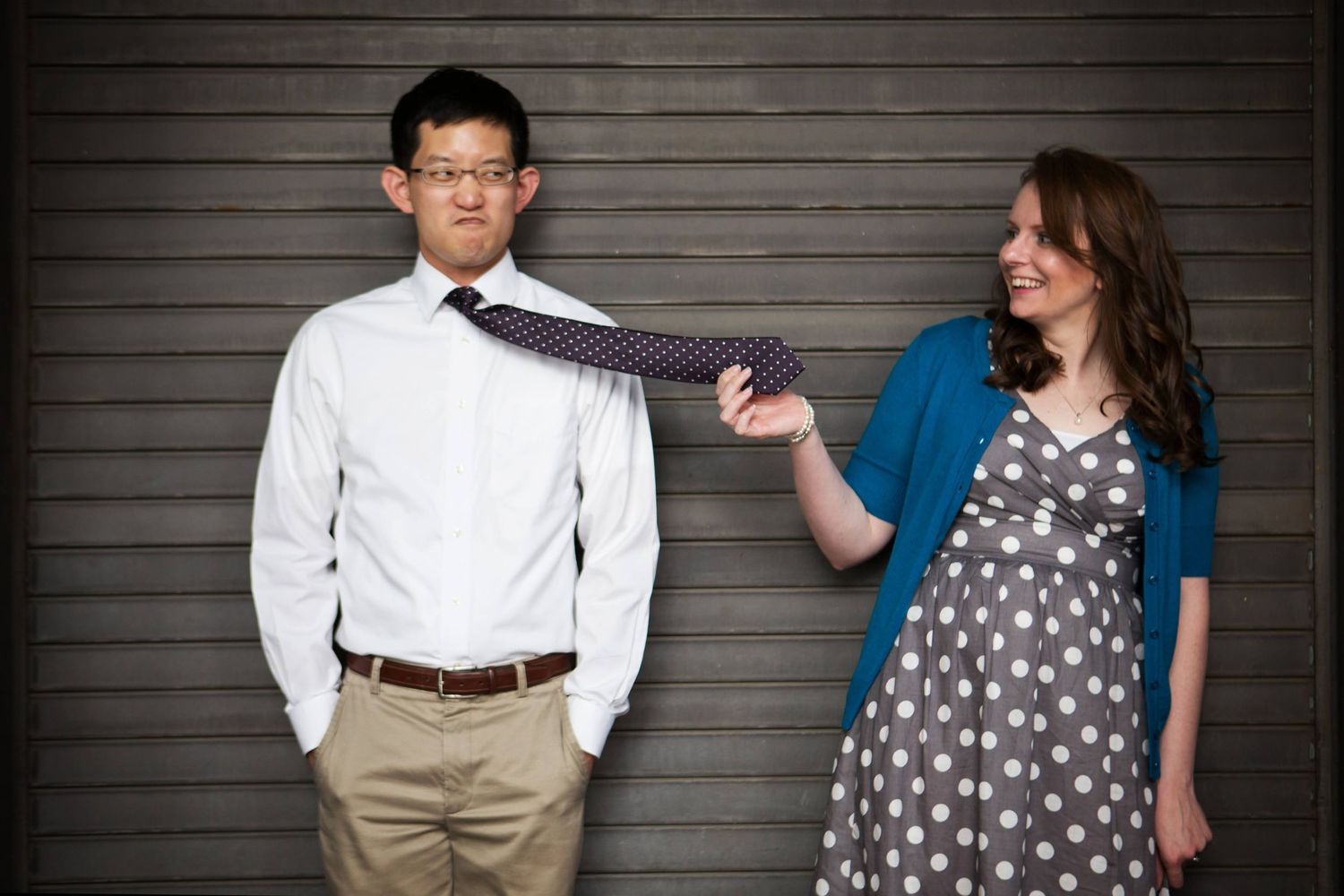 Playful engagement photo with couple in casual attire against gray wall.