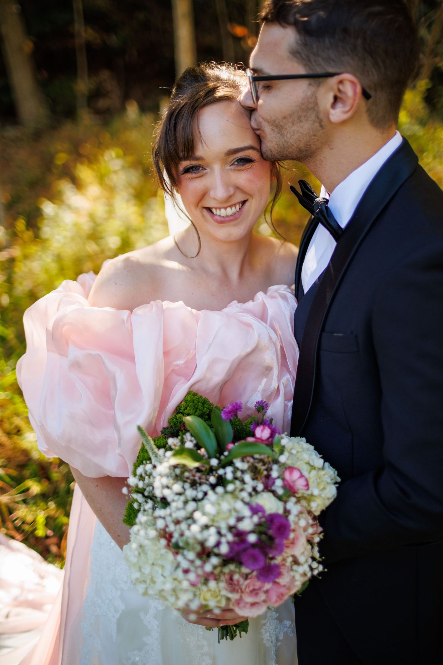 Smiling bride in pink off-shoulder gown holds bouquet as groom in tuxedo kisses her cheek outdoors.