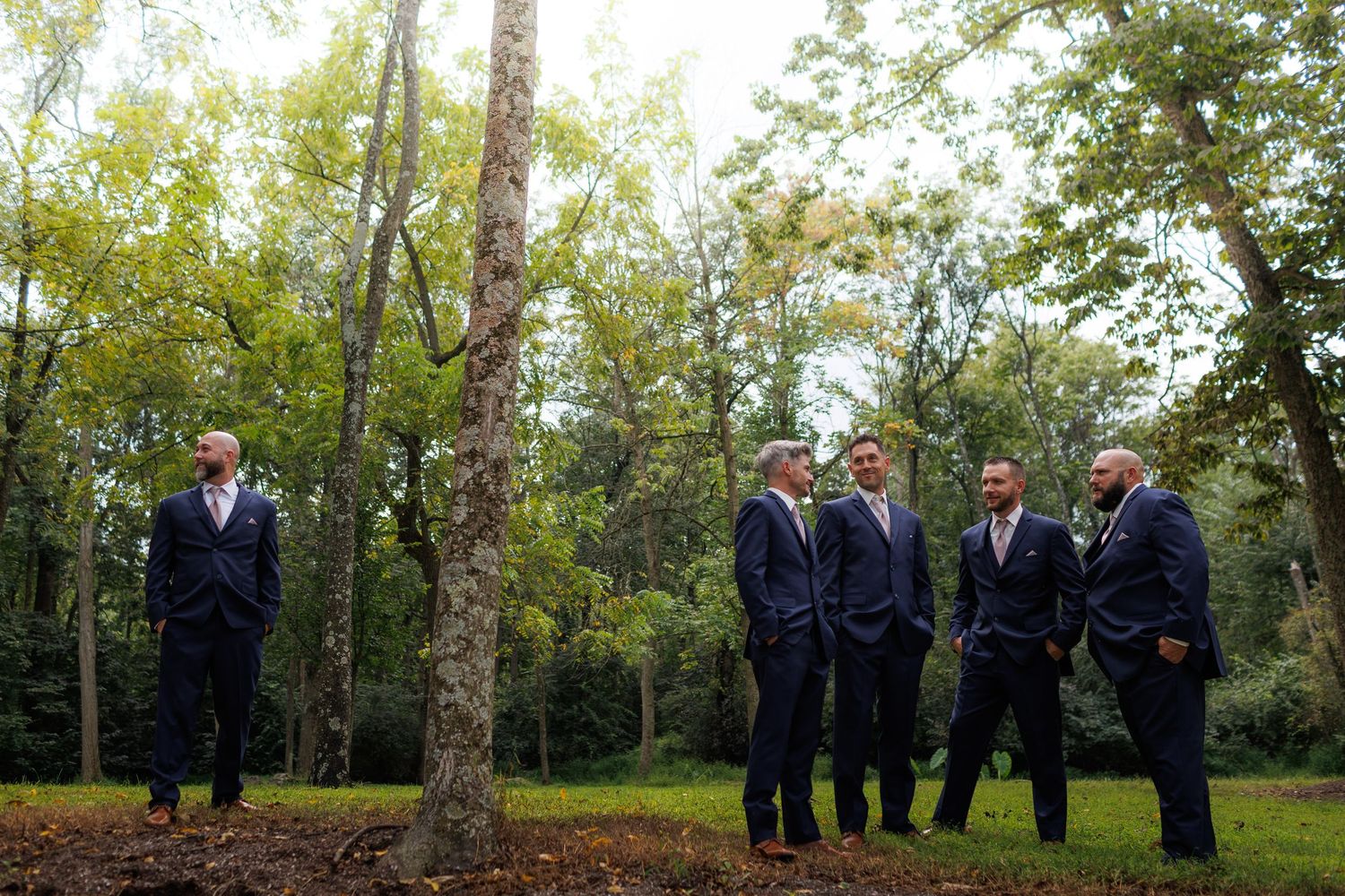 Wedding party in dark suits standing in a line against a wooded outdoor backdrop.