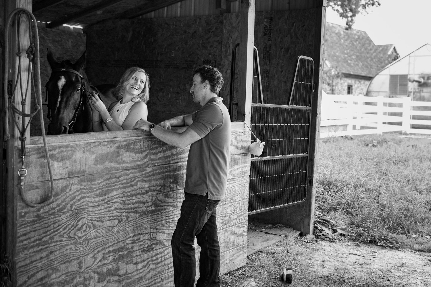 A man and woman smile at each other beside a horse stable in a black and white farm photo.