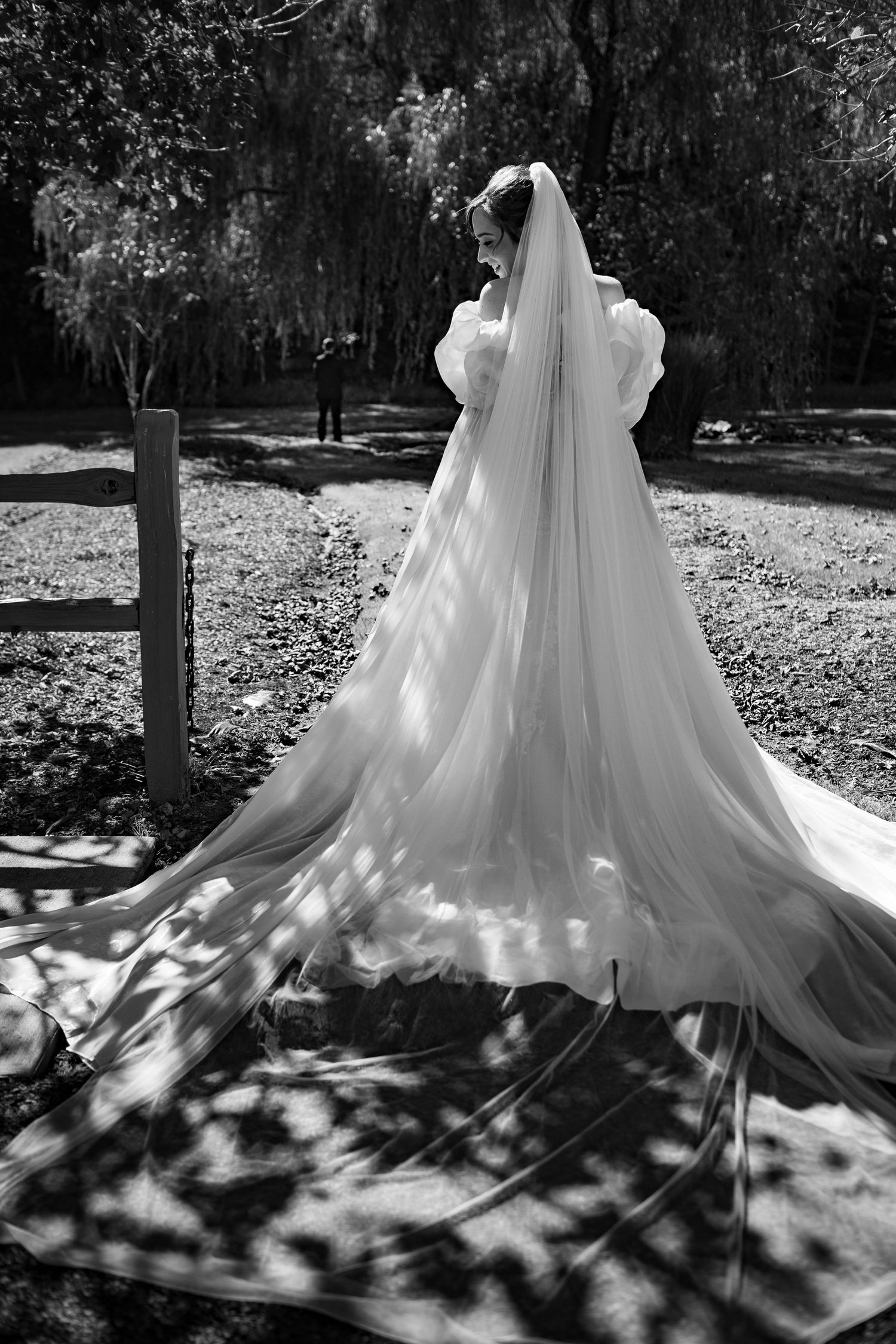 Bride in elegant white gown with long flowing veil stands in sunlit park, viewed from behind.
