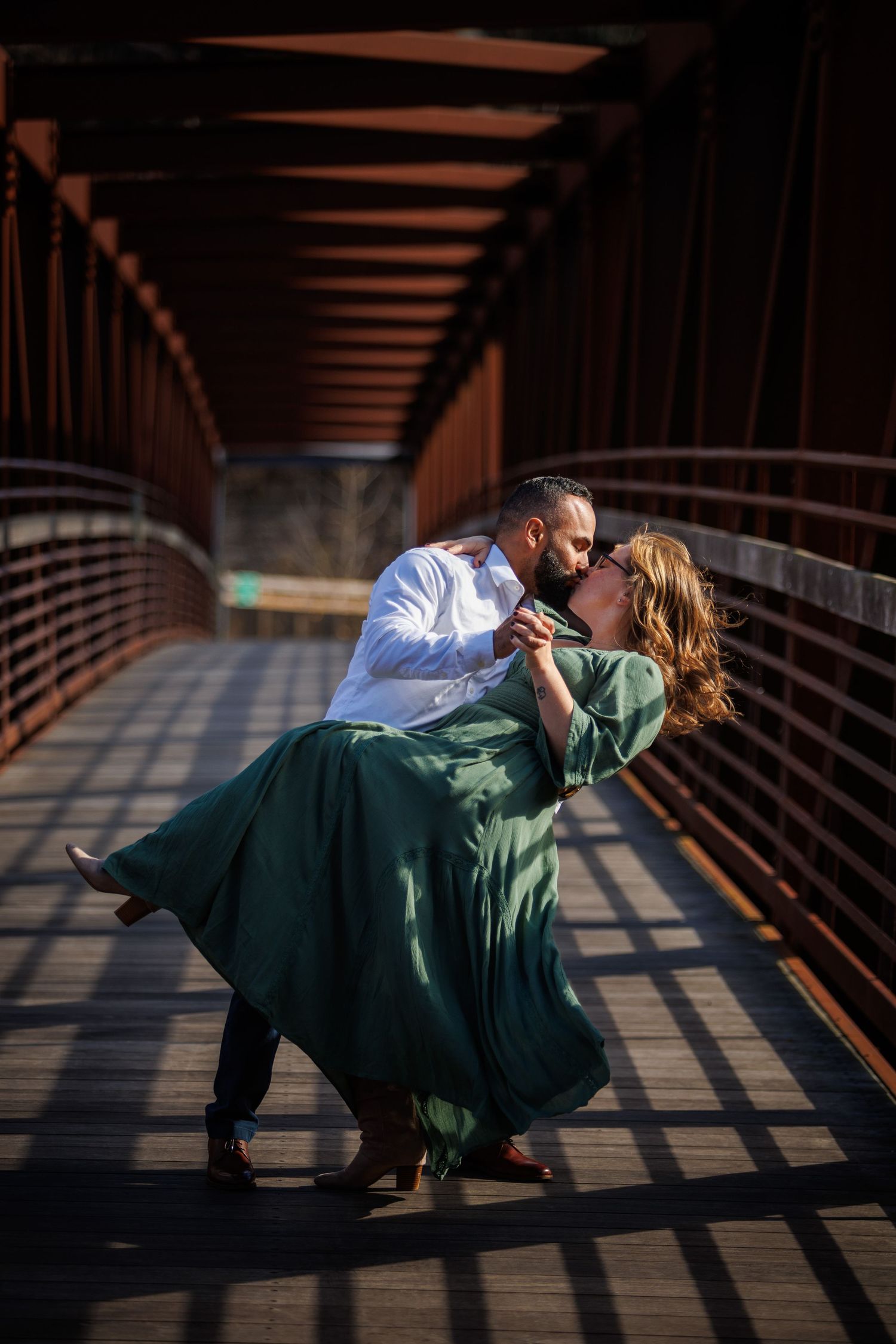 A romantic couple in formal attire share a dramatic dip pose on a wooden bridge during a sunset photo session.