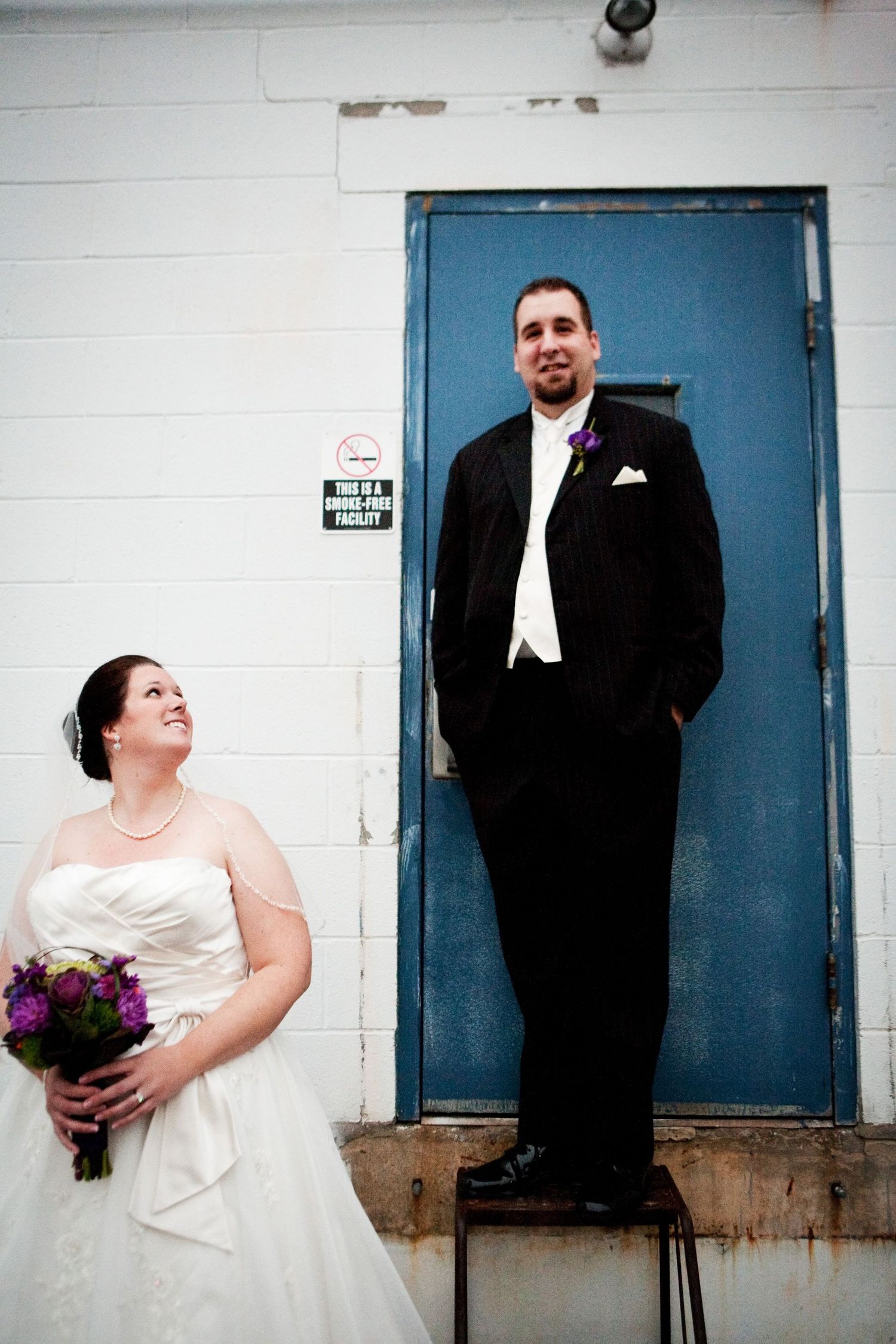 A bride and groom pose playfully in front of a blue door, with her holding a purple bouquet and him standing elevated.