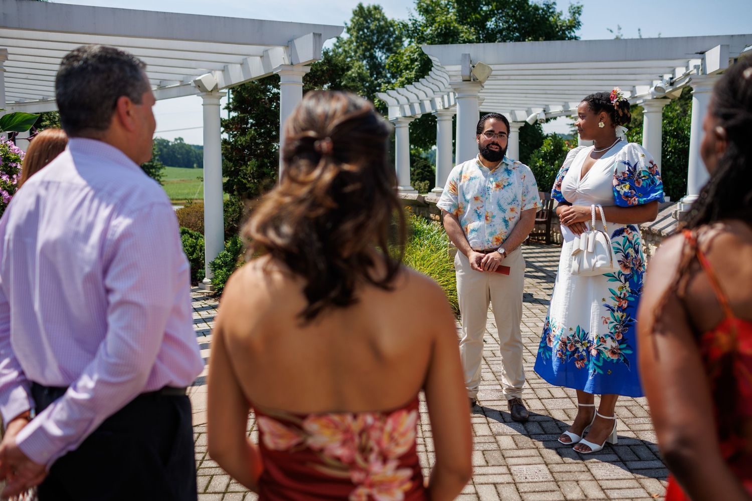 Outdoor wedding ceremony with guests gathered under a white pergola surrounded by lush greenery on a sunny day.