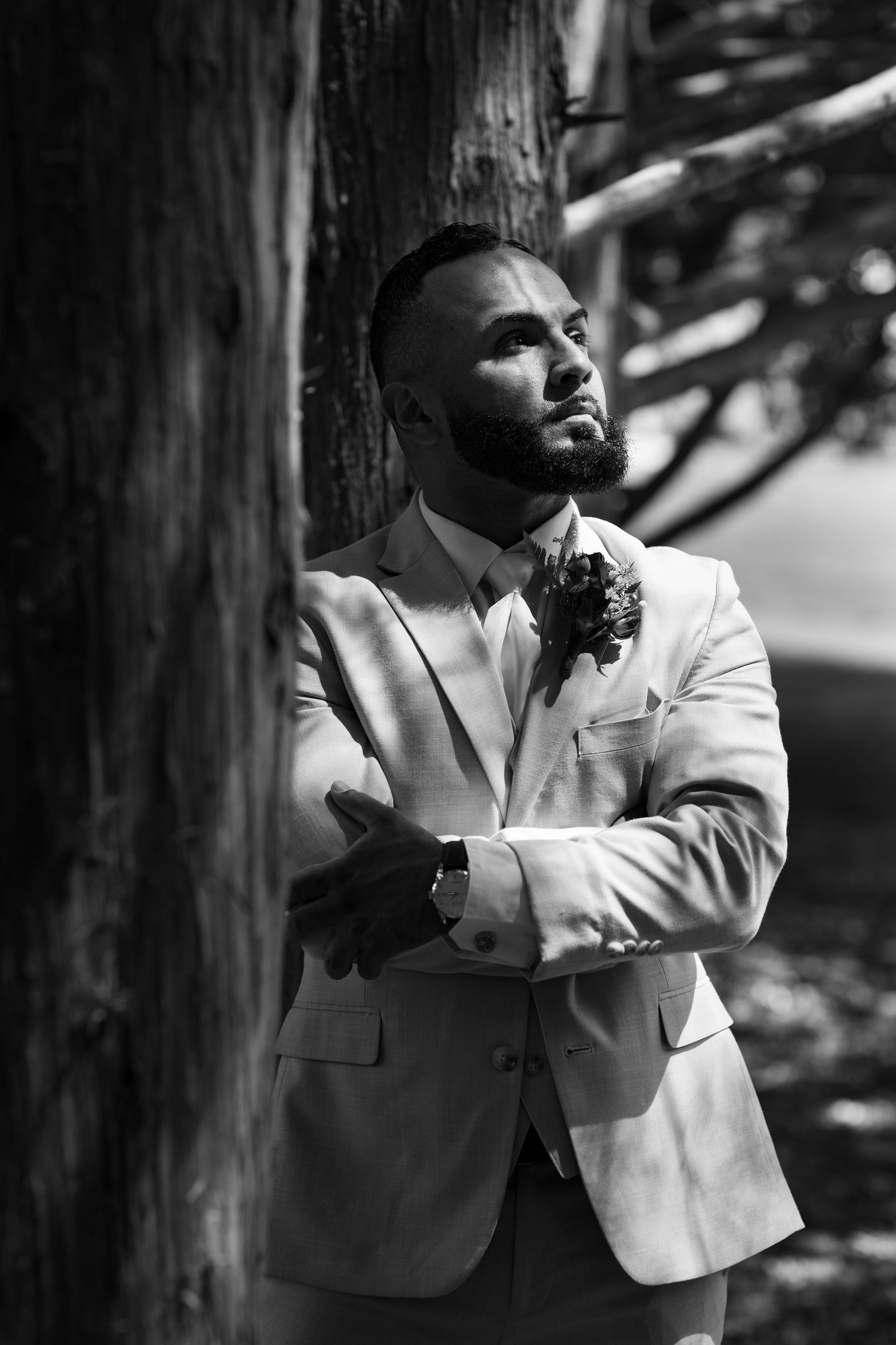 Elegant groom in light suit with boutonniere leaning against tree in black and white wedding portrait.