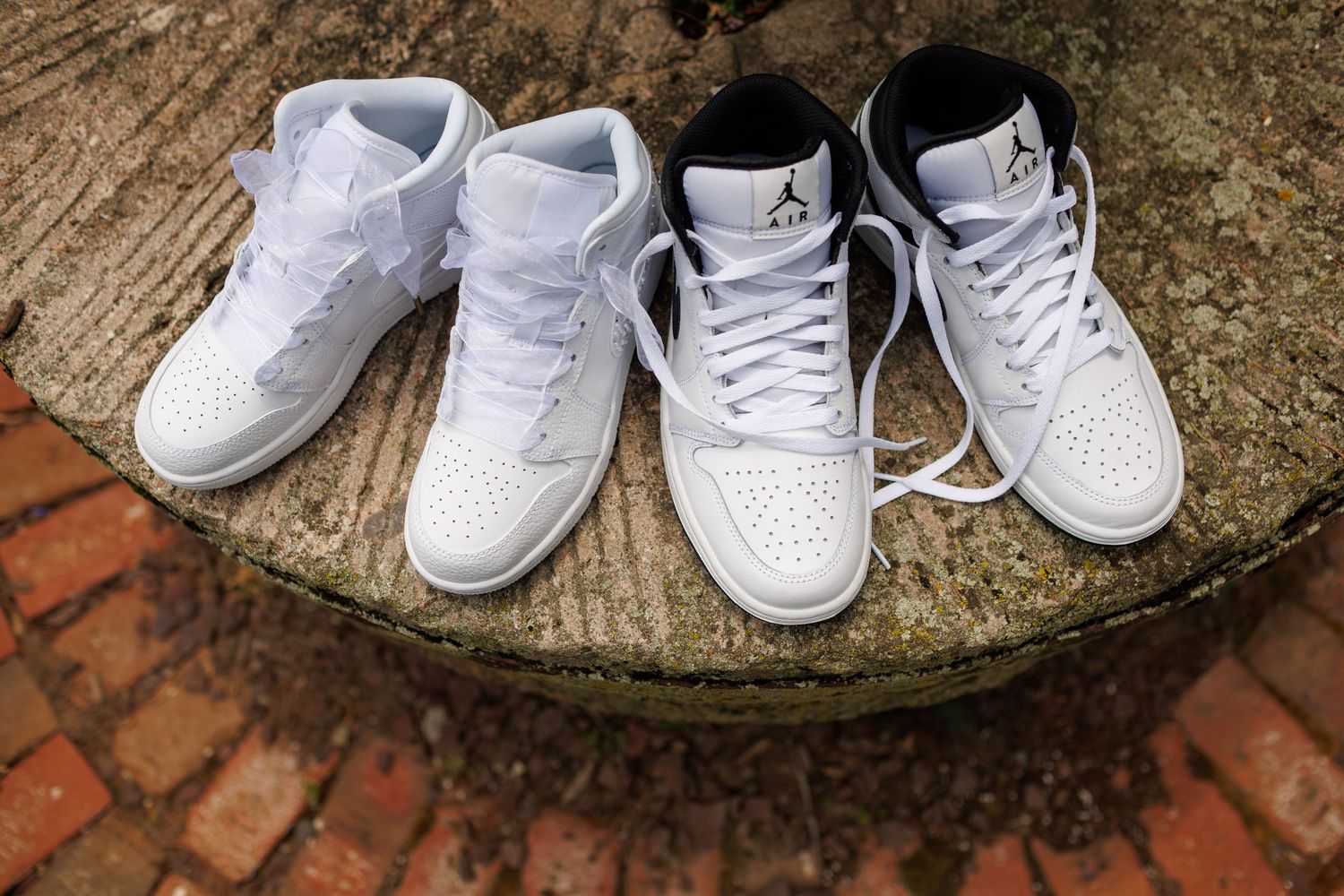 Two pairs of white Air Jordan 1 Mid sneakers displayed on a stone surface with brick background.