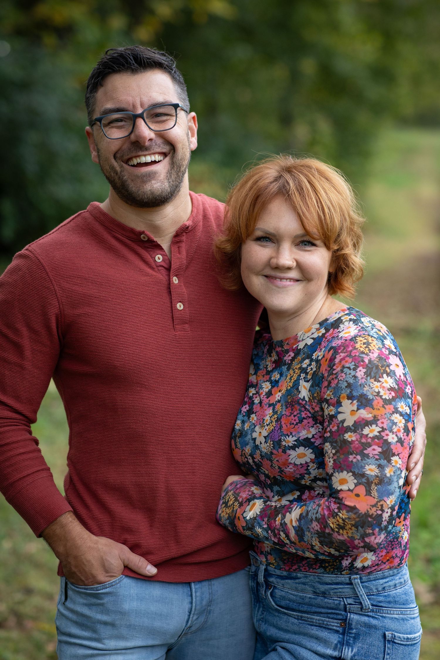 A couple poses together outdoors wearing casual fall attire.