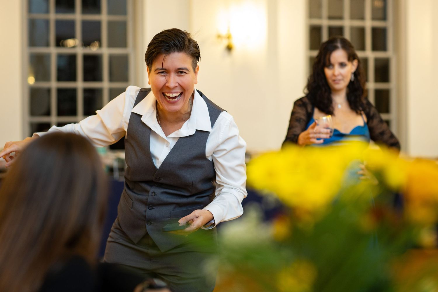 Warm indoor gathering with yellow flowers in foreground and people socializing in background.