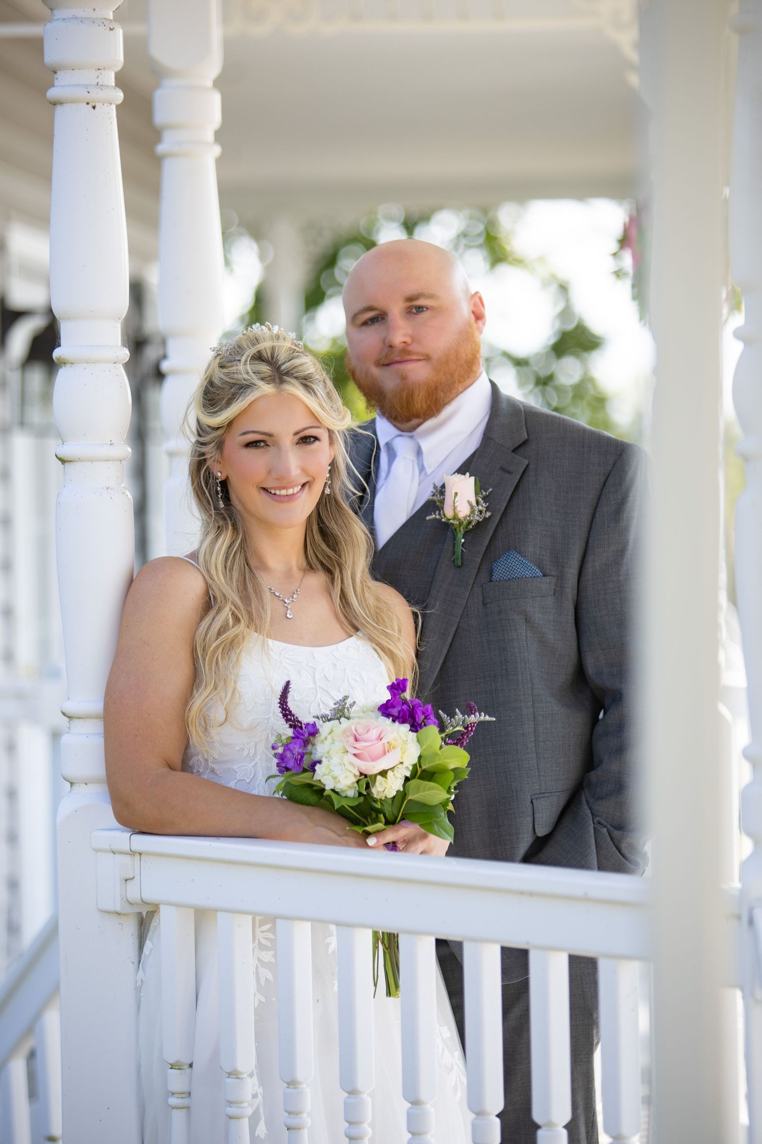 A happy couple poses for a wedding portrait on a white porch with bride holding a purple and white floral bouquet.
