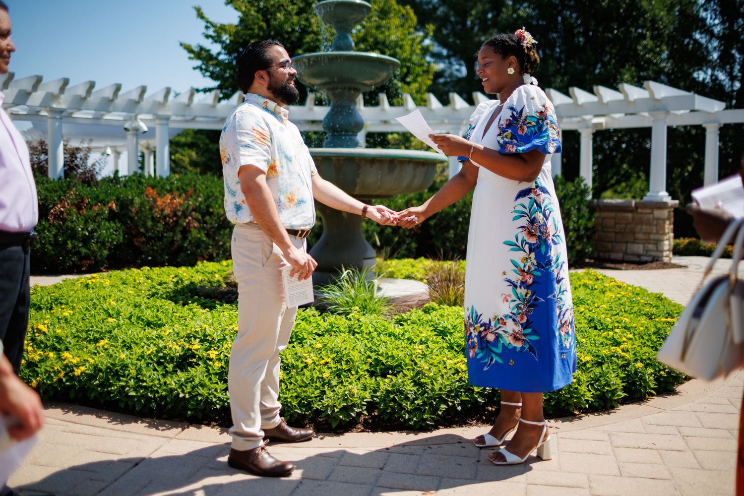 Couple exchanging vows in a sunny garden ceremony near a fountain, surrounded by lush greenery and white arbor.