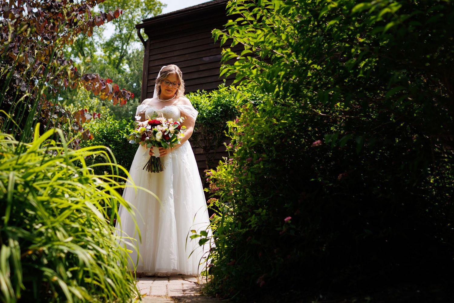 Bride in white wedding gown holding a bouquet walking through a green garden pathway.
