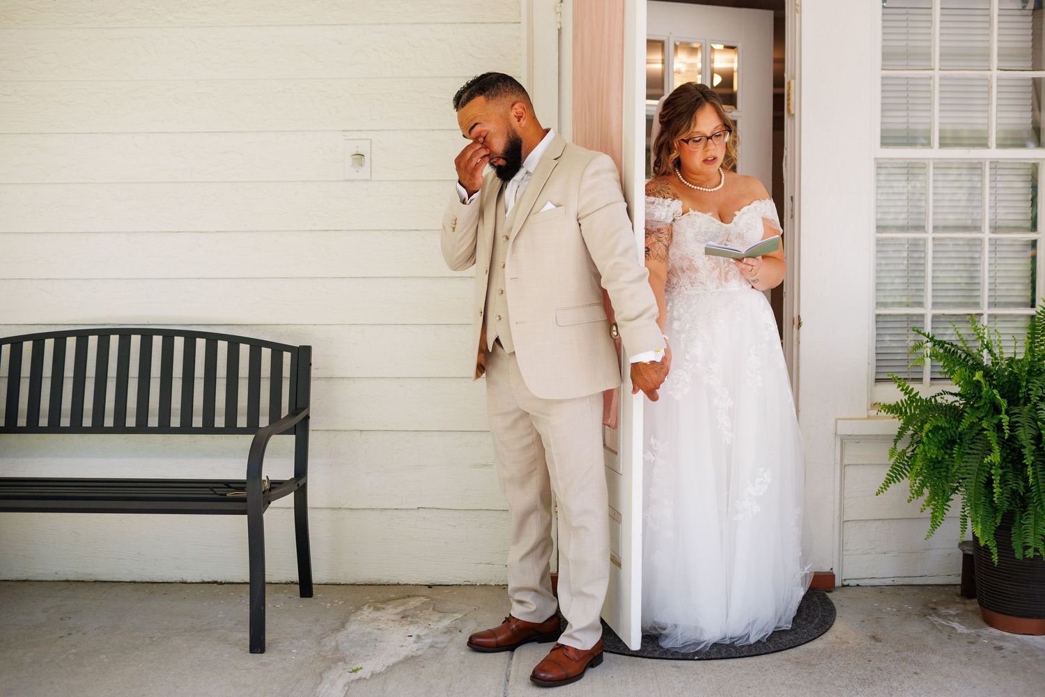 Emotional groom wipes tears during first look with bride in lace wedding gown at doorway.