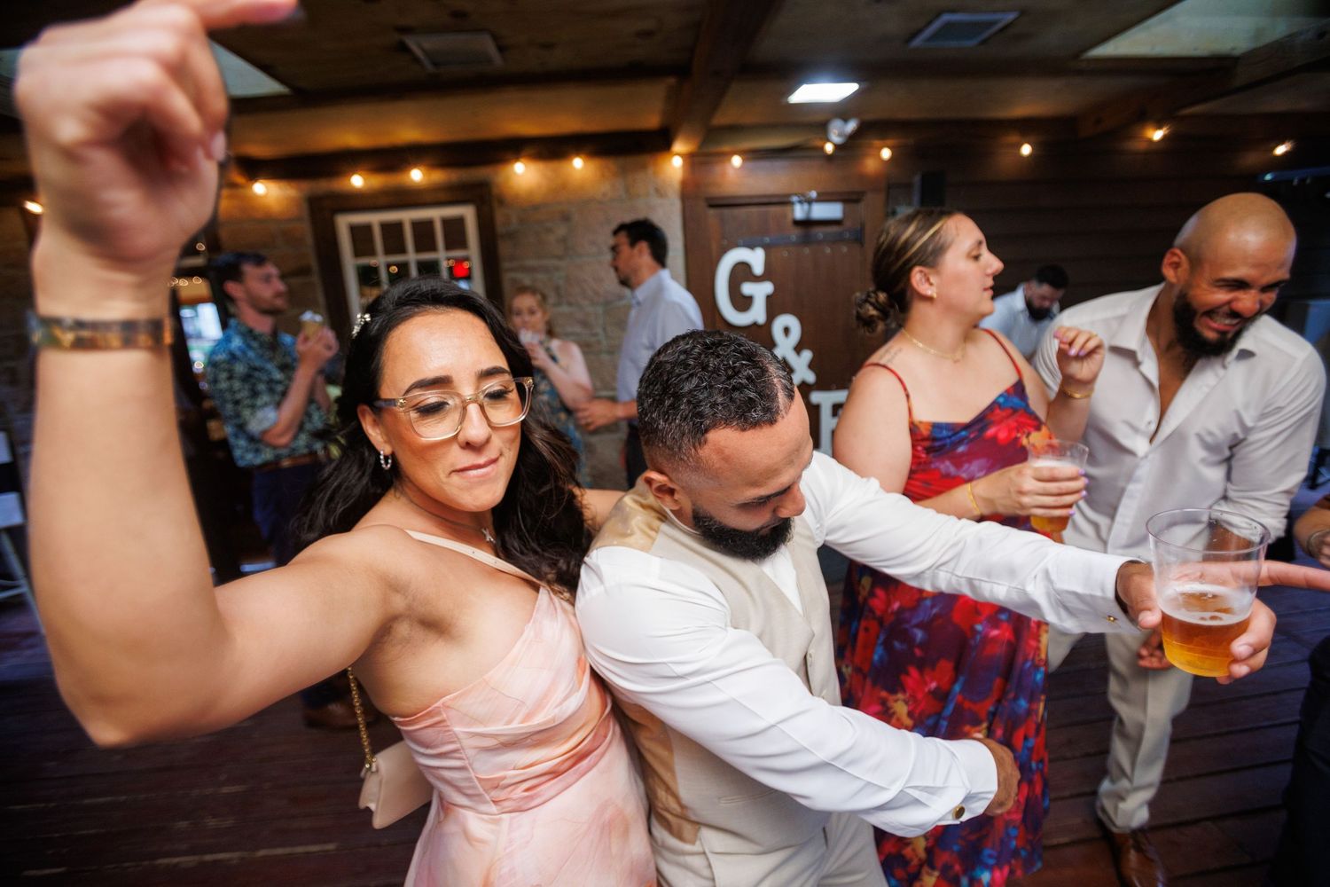 Guests dance and celebrate joyfully at indoor wedding reception under warm string lights.