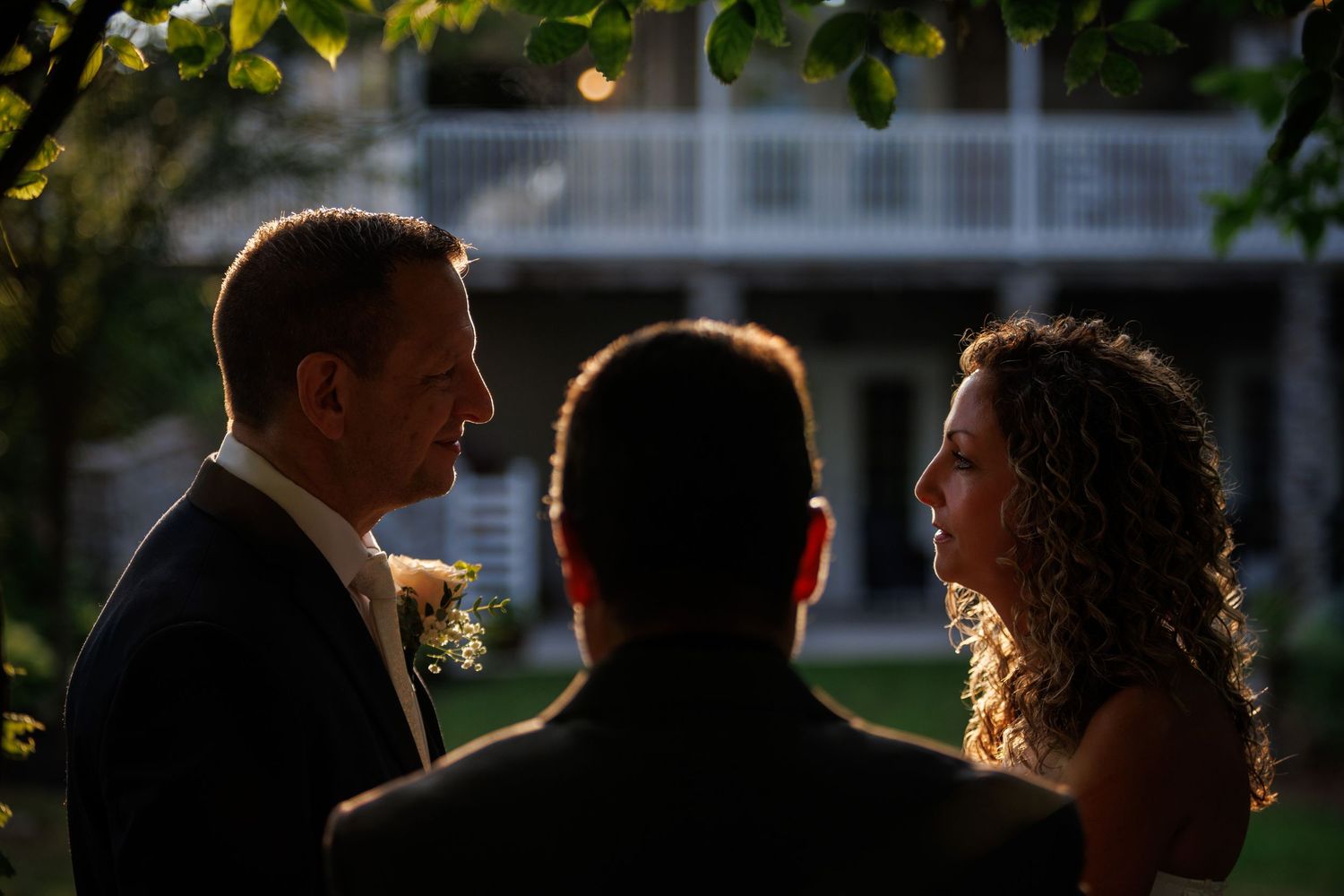 Couple exchanging vows outdoors at golden hour with officiant, elegant white house backdrop, warm sunlight highlighting bride and groom.