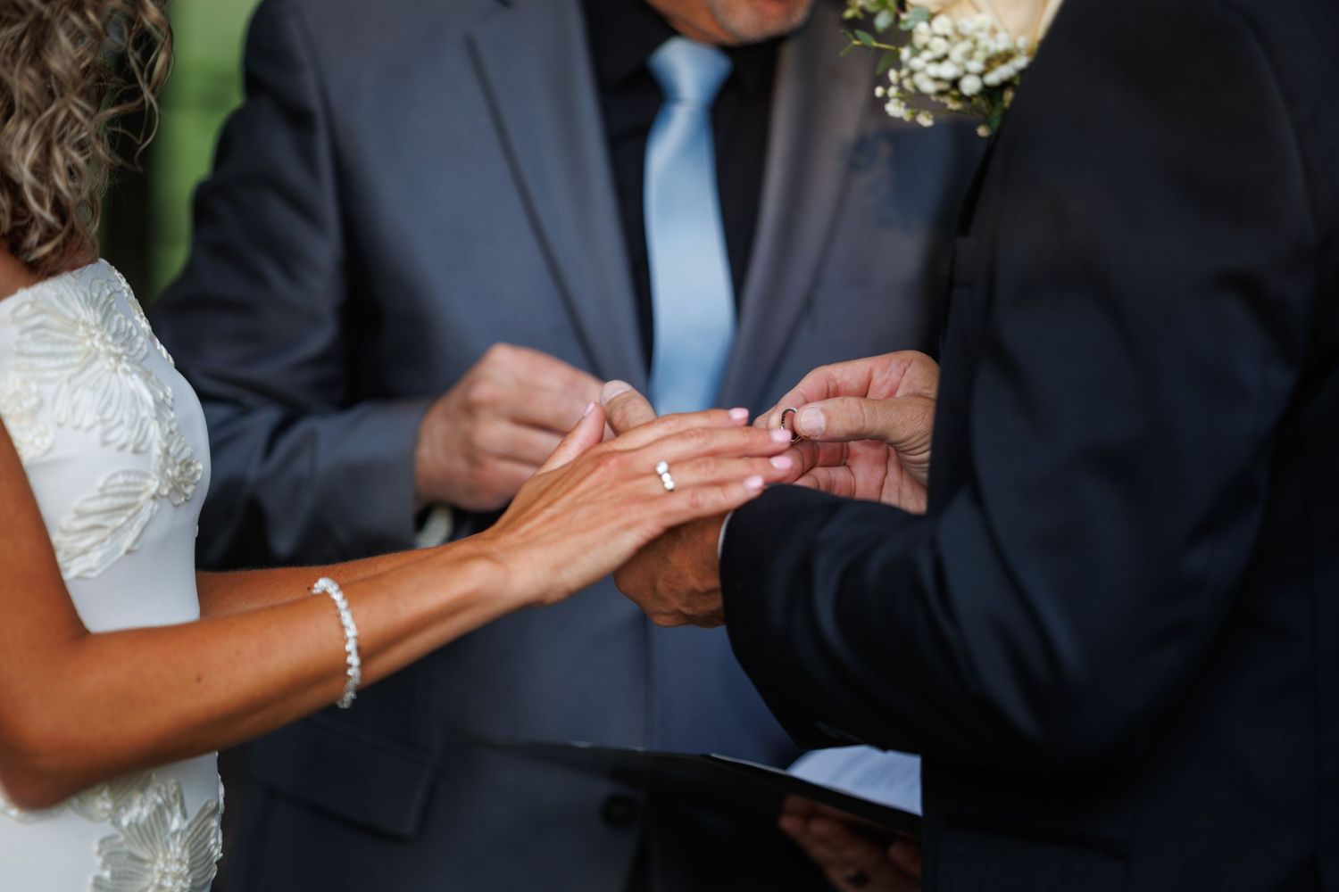 Groom placing a wedding ring on bride's finger during a ceremony, with an officiant holding a book in the background.