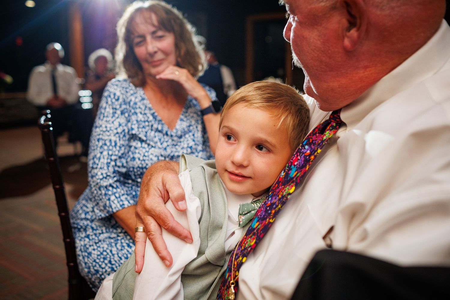A young child sits on an older mans lap while a woman smiles warmly at a wedding.