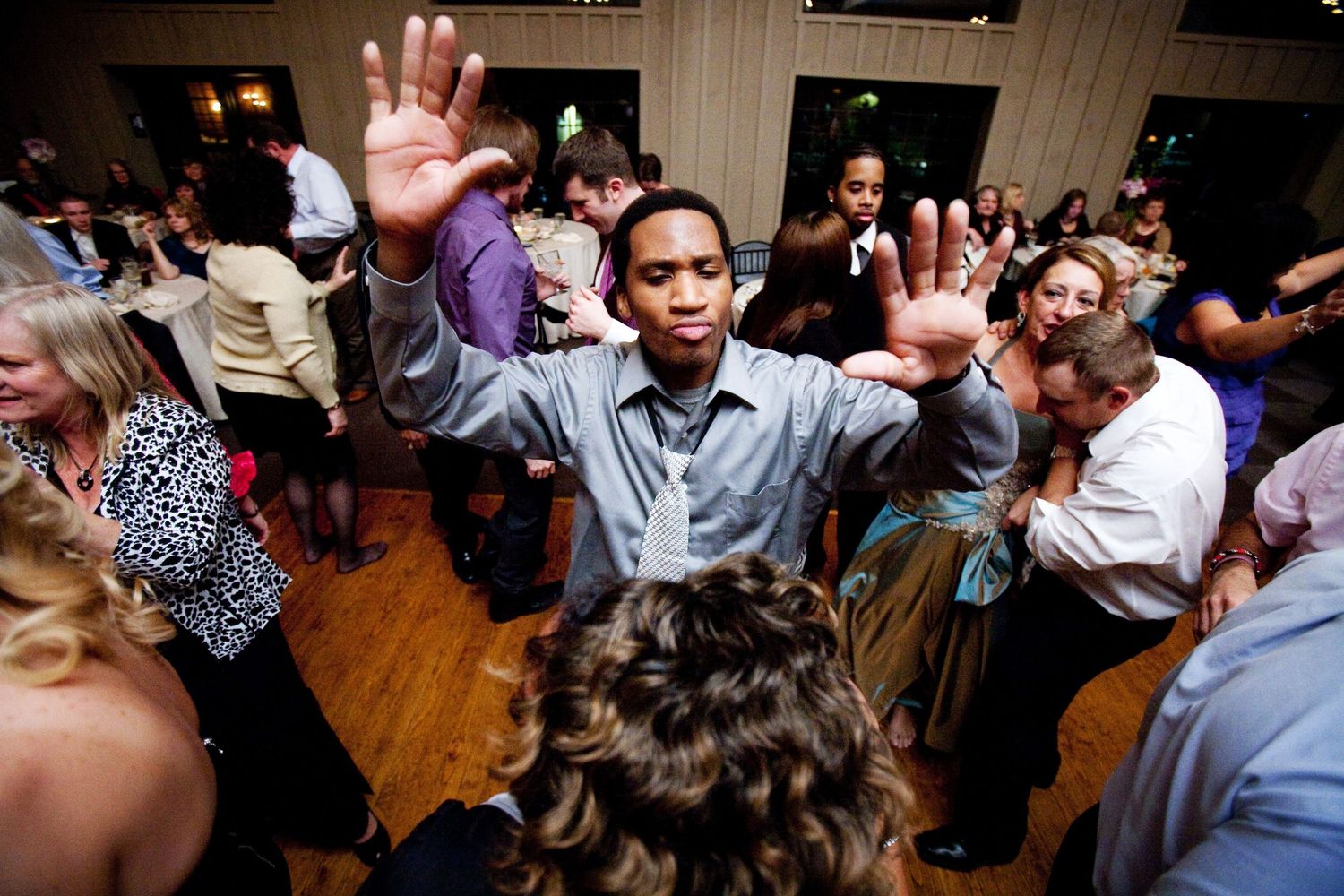 People with raised hands dance and celebrate together on a wooden floor at what appears to be a party or reception event.