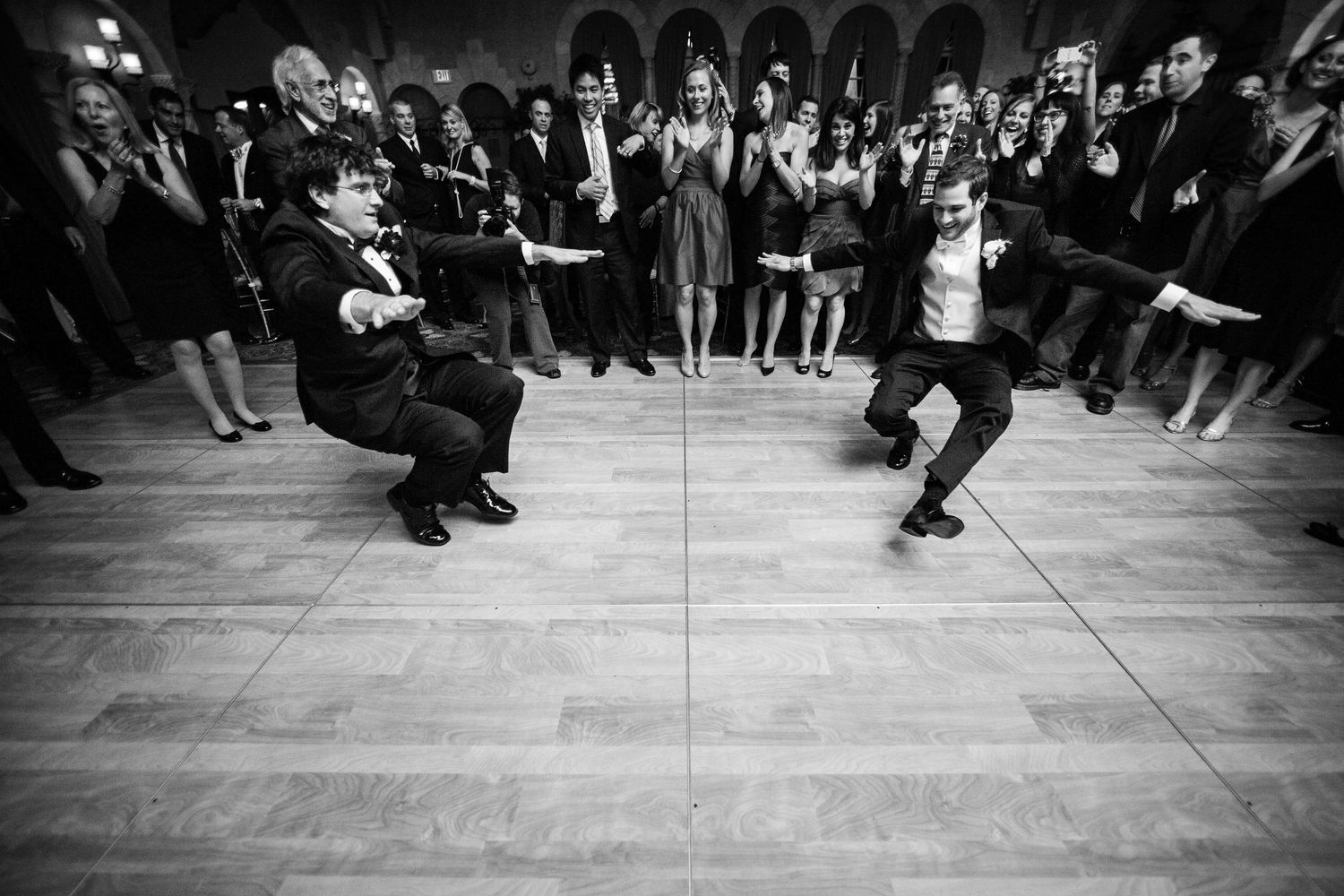 Two people engage in an energetic dance battle on a wooden floor surrounded by a crowd of spectators in black and white.