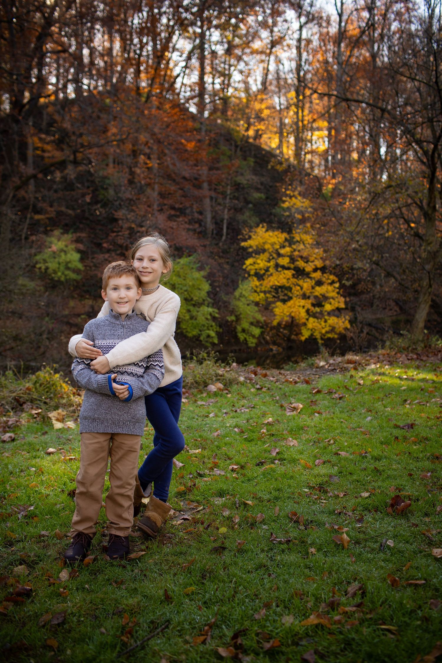Playful interaction between two people on a grassy hill surrounded by fall foliage.