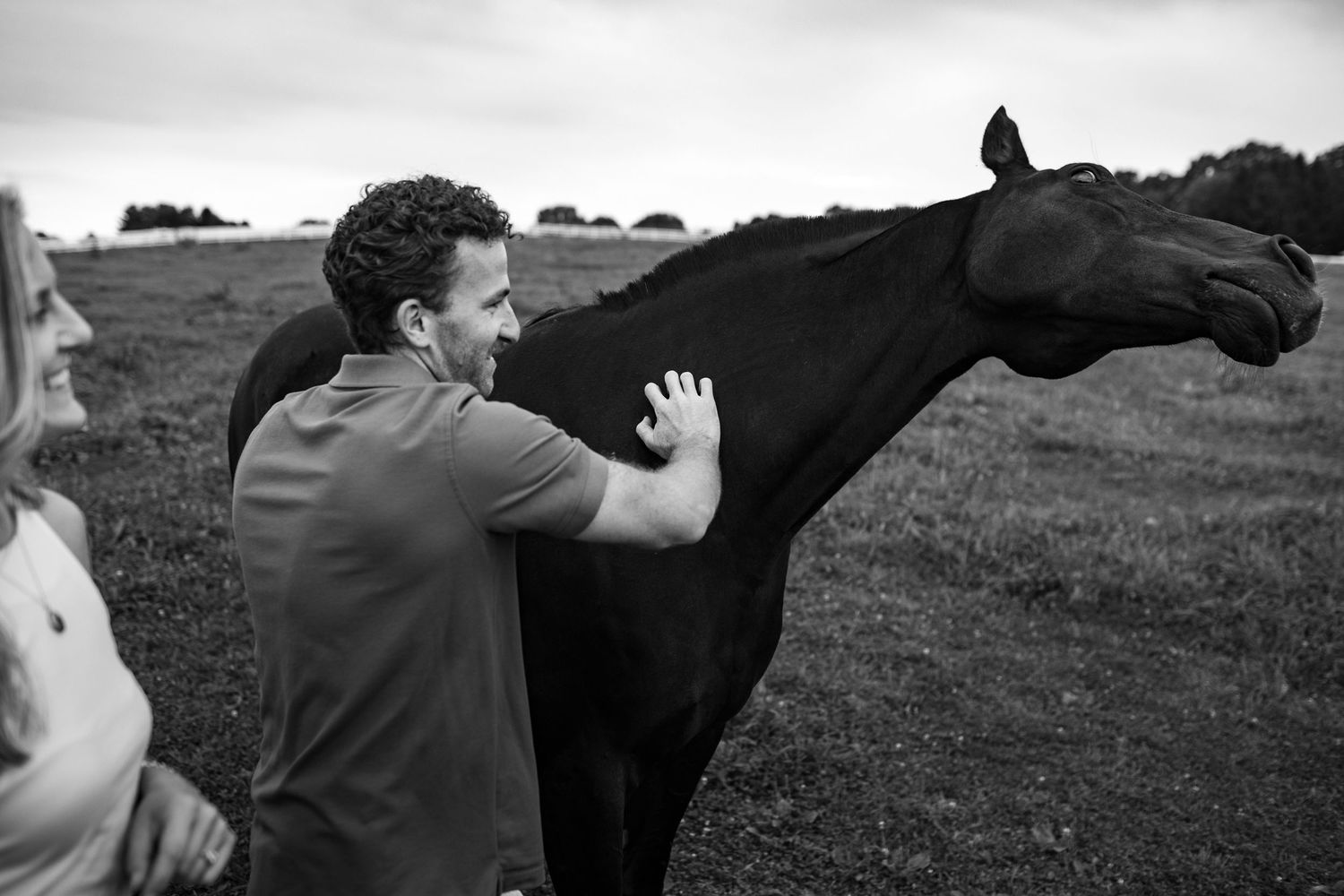 Man petting a dark horse in a field while a woman stands nearby in a black and white outdoor photo.