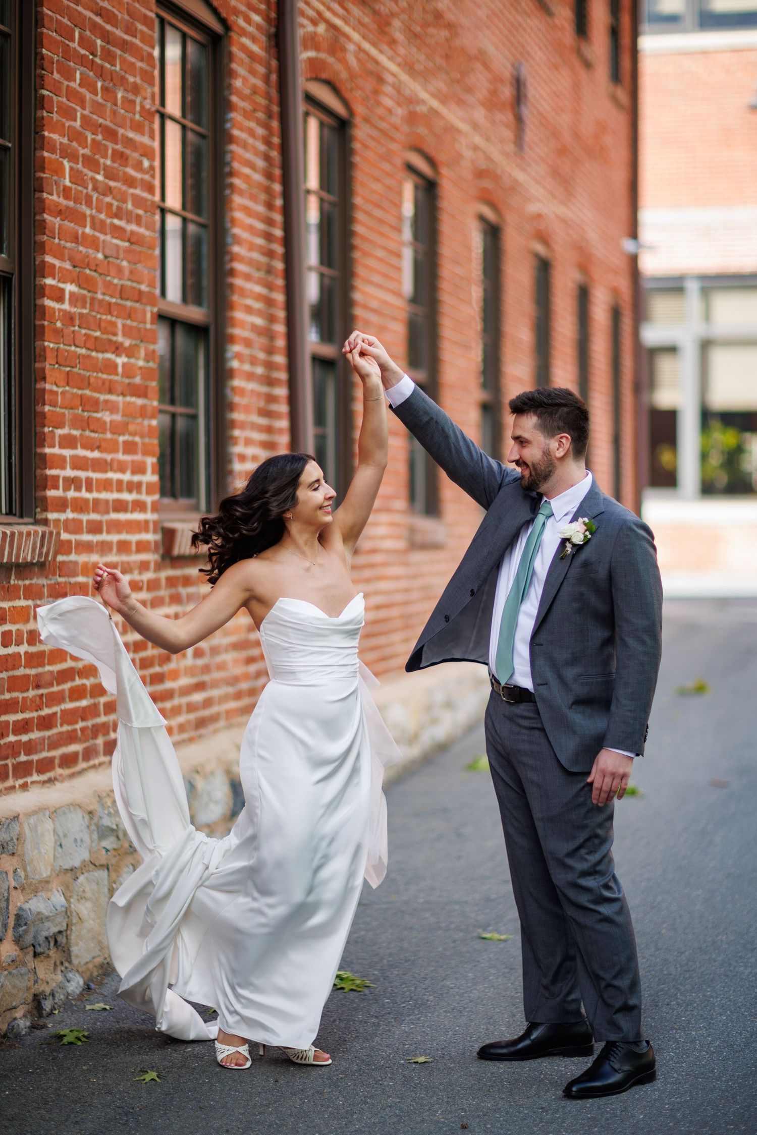Bride and groom dancing joyfully in an alley beside a historic red brick building on their wedding day.