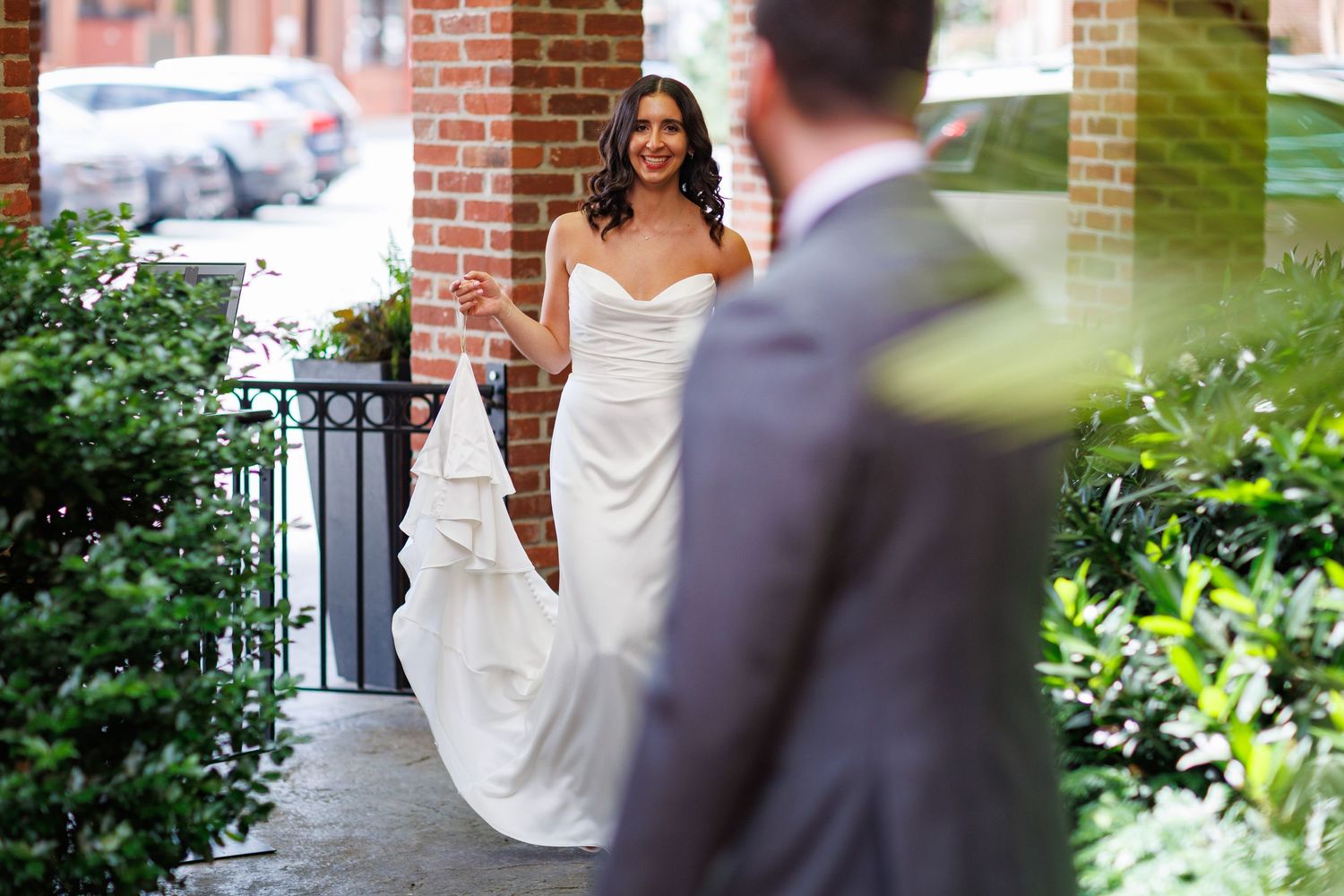 Bride in elegant white strapless gown holding train, smiling at groom during romantic first look outside brick building.