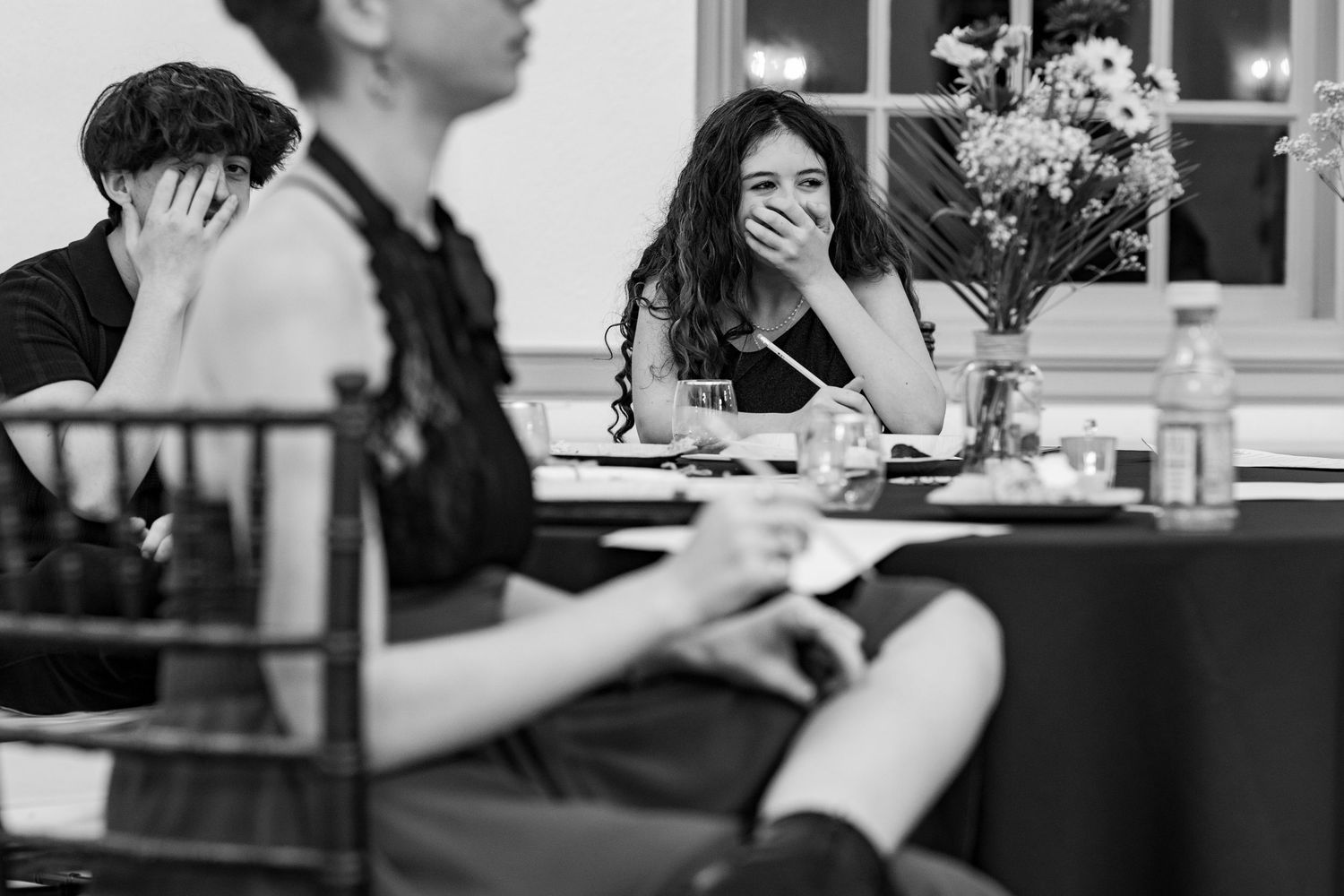 People seated at tables during formal event with window lighting.