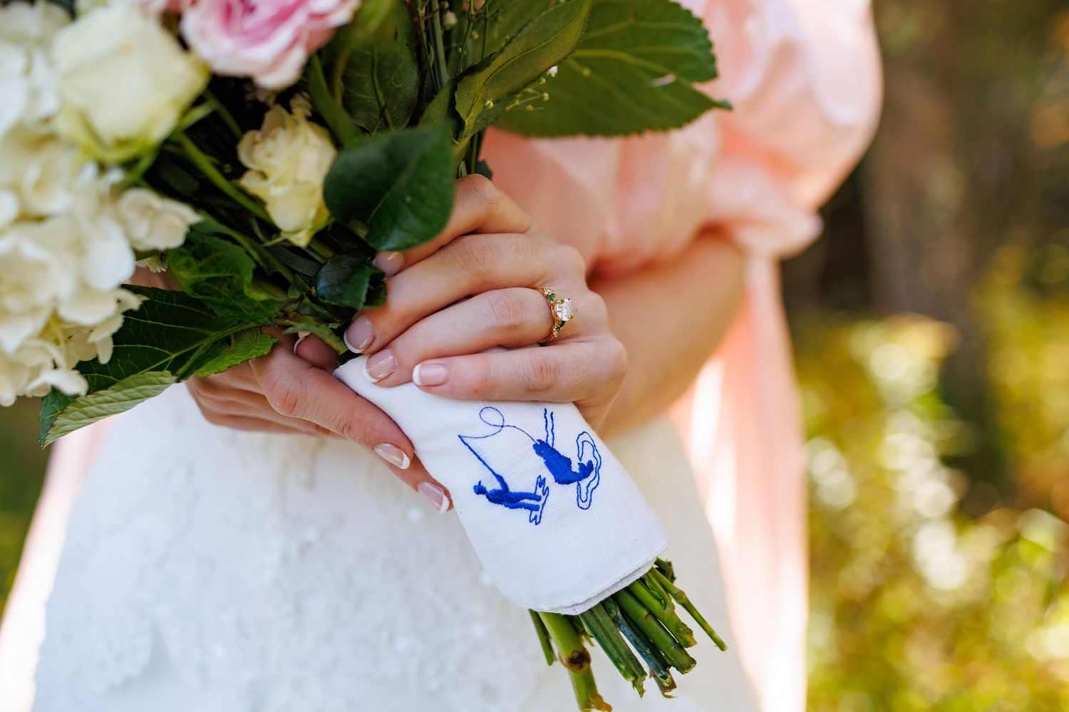 Bride holding a floral bouquet with a monogrammed handkerchief, wearing a diamond ring, in a white wedding dress.