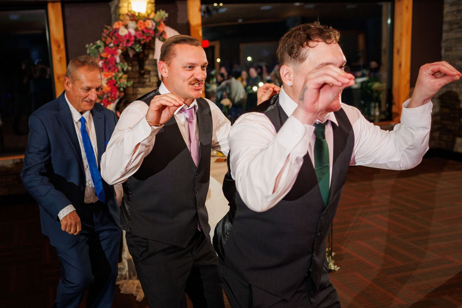 Two groomsmen in vests dance energetically and make funny faces at a wedding reception.