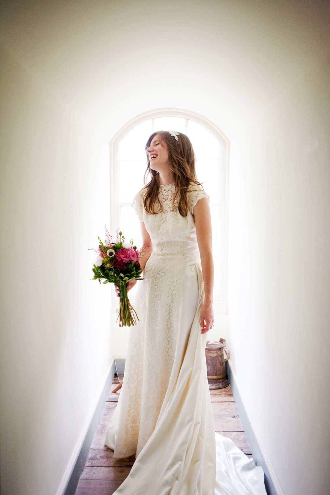 Smiling bride in white lace gown holding pink bouquet stands in arched window light on wedding day.