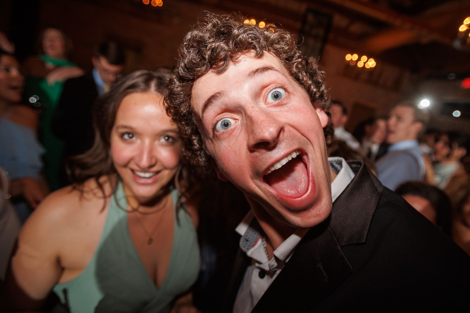 Excited young man making a funny face at a party with a smiling woman in a green dress behind him.
