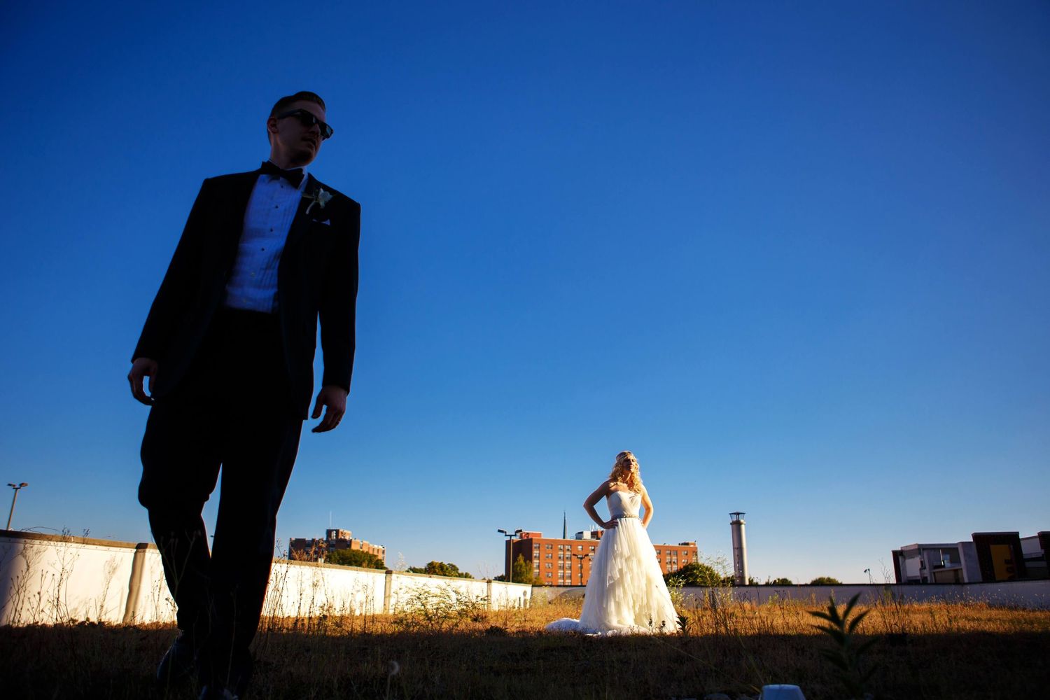 Silhouetted figures in formal wear against an industrial sunset backdrop.
