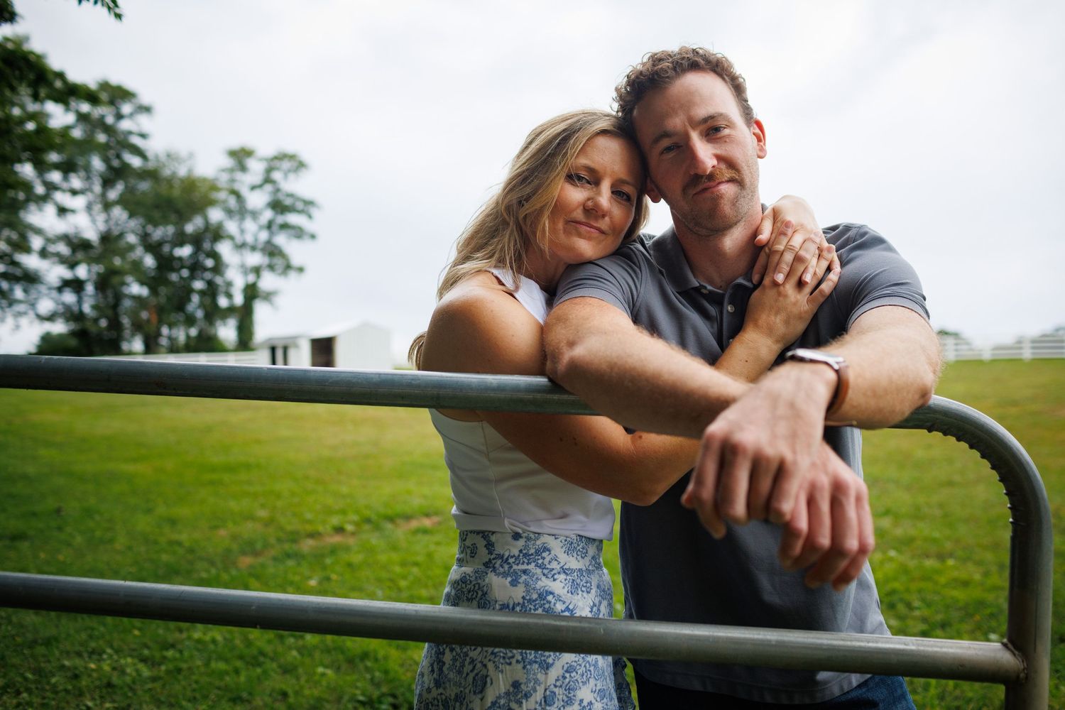 A smiling couple leans on a metal gate in a lush green field, posing for an outdoor engagement photo.
