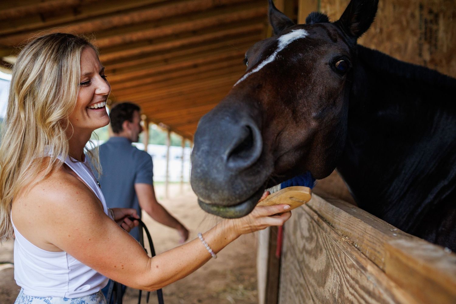 Smiling blonde woman petting a dark horse in a stable, enjoying a friendly interaction at an equestrian facility.