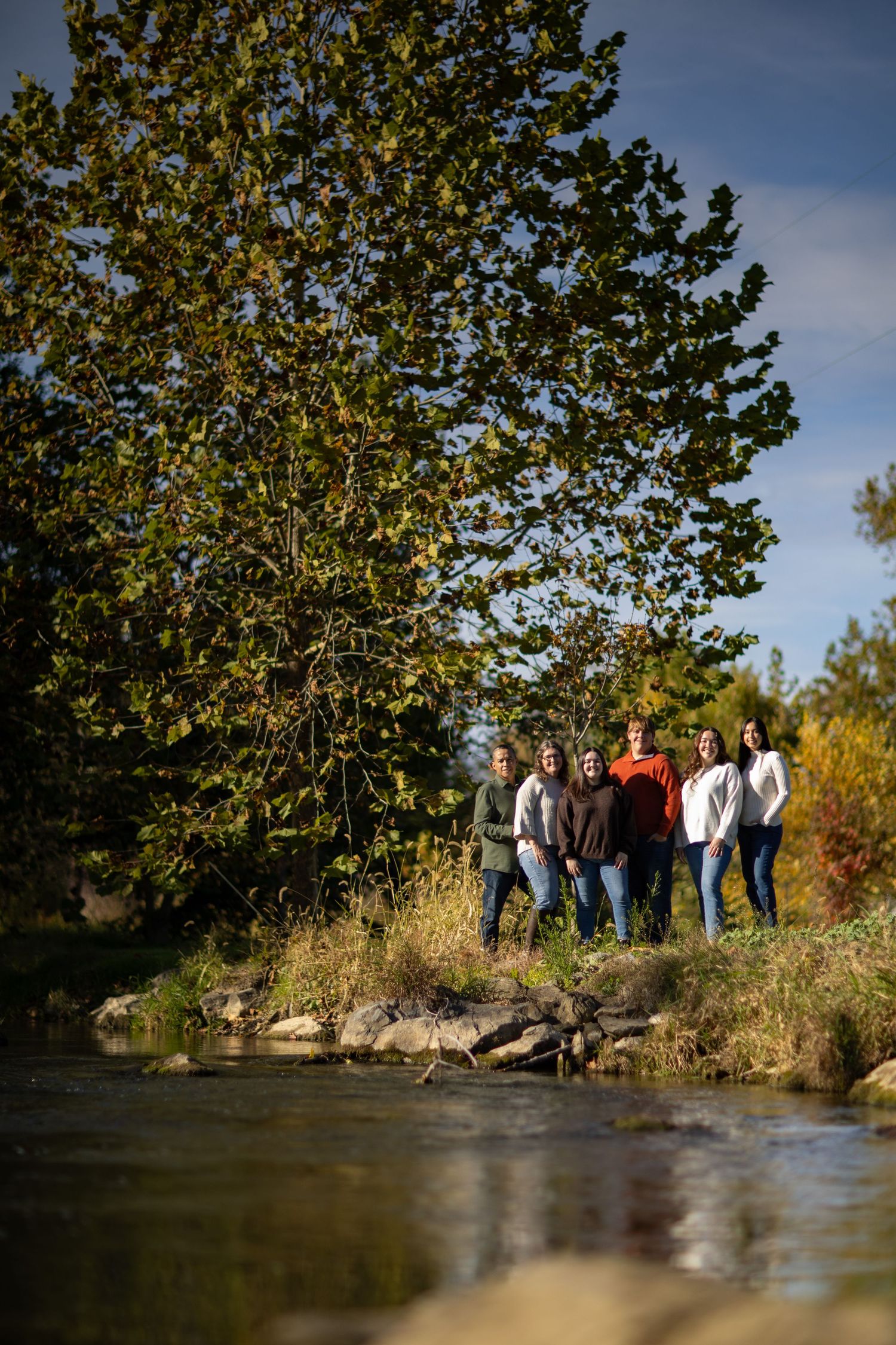 A group stands together on a riverbank surrounded by autumn trees and foliage on a sunny day.