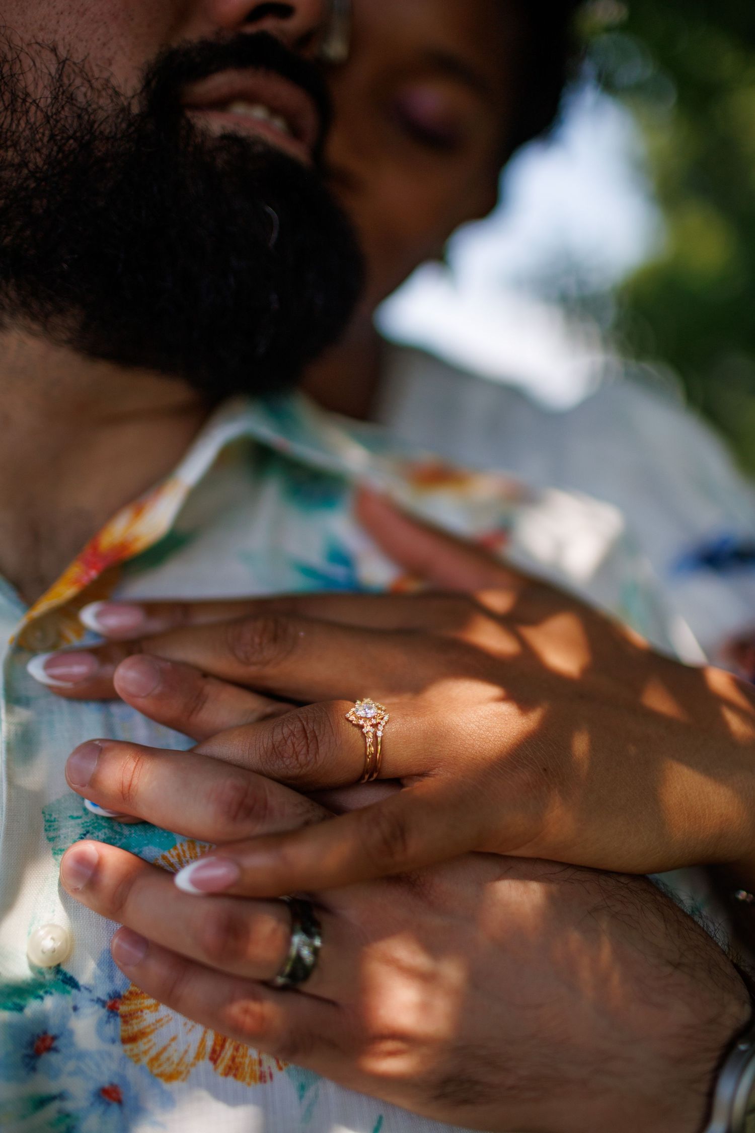 Close-up of couple embracing, showcasing a gold diamond engagement ring and wedding band in dappled sunlight.